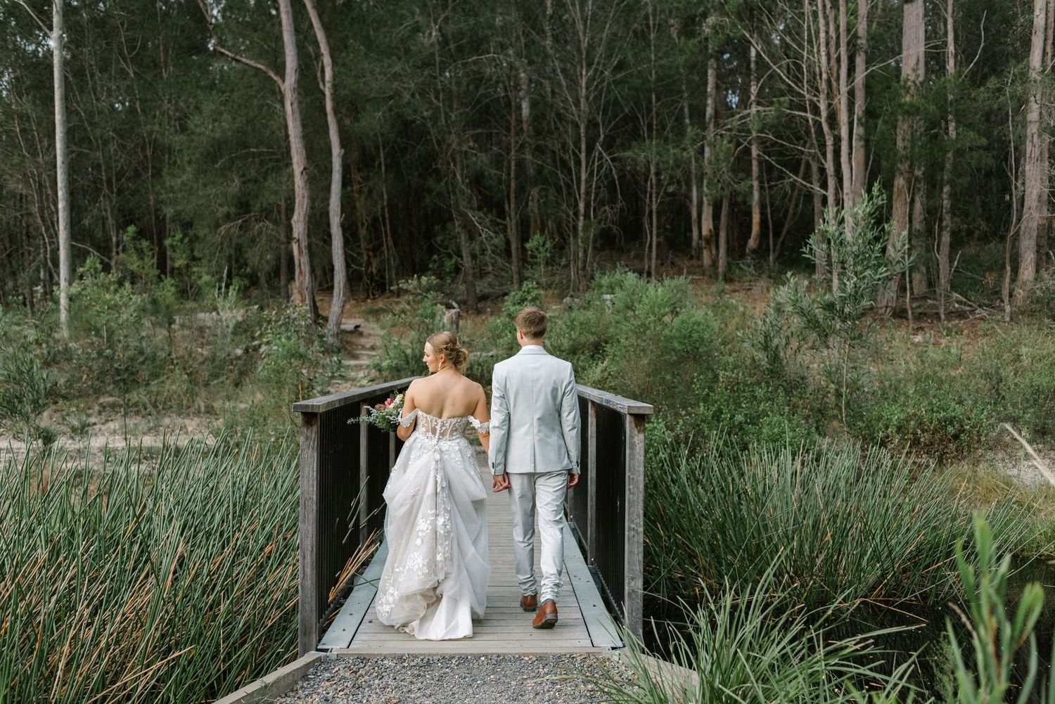 Bride and groom walk hand-in-hand across a wooden bridge surrounded by lush greenery and tall trees.