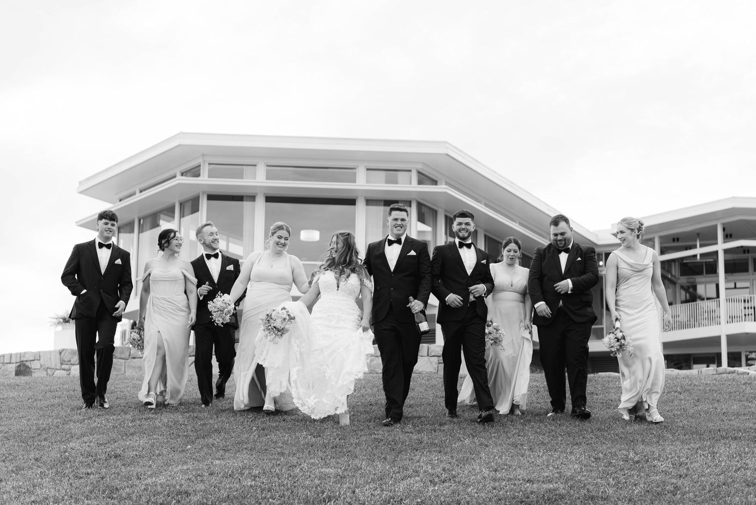 Large wedding party posing in formal attire on a sprawling lawn with buildings in background.