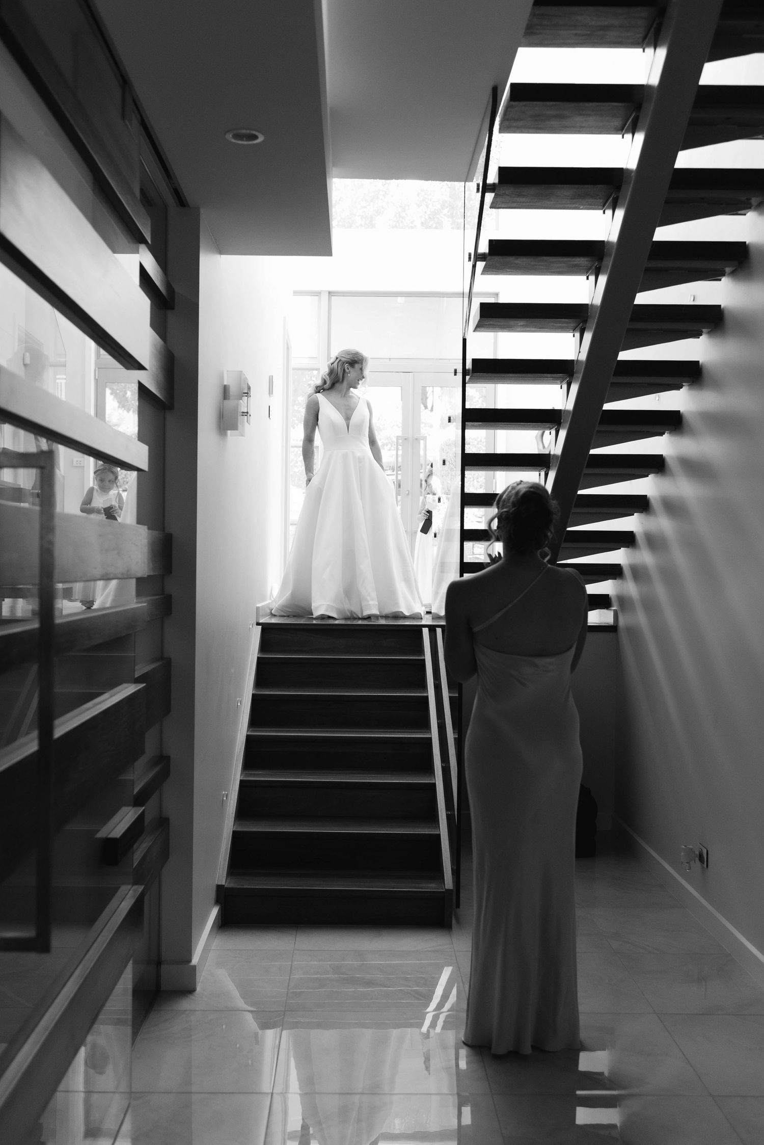 Black and white photo of a bride in a white gown on stairs while a bridesmaid looks up at her.