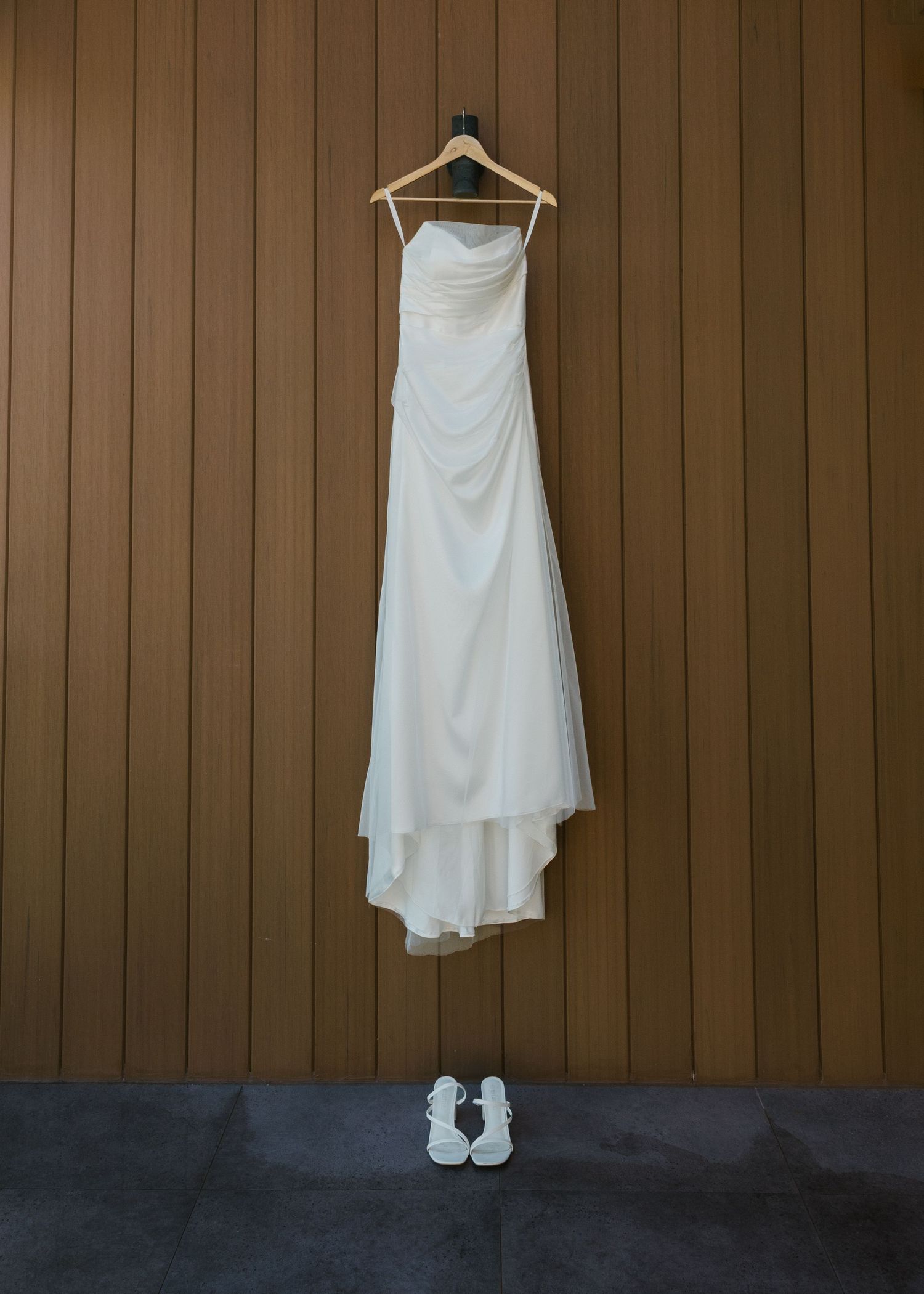 White strapless wedding gown hanging on wooden wall with white bridal sandals placed neatly below.