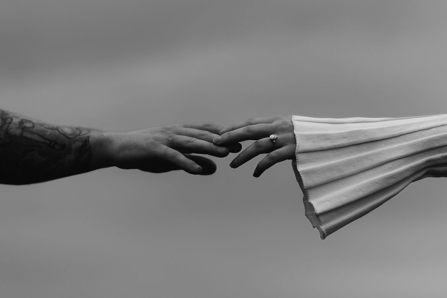 Black and white photo of two hands reaching toward each other, one tattooed, the other holding a flowing fabric.