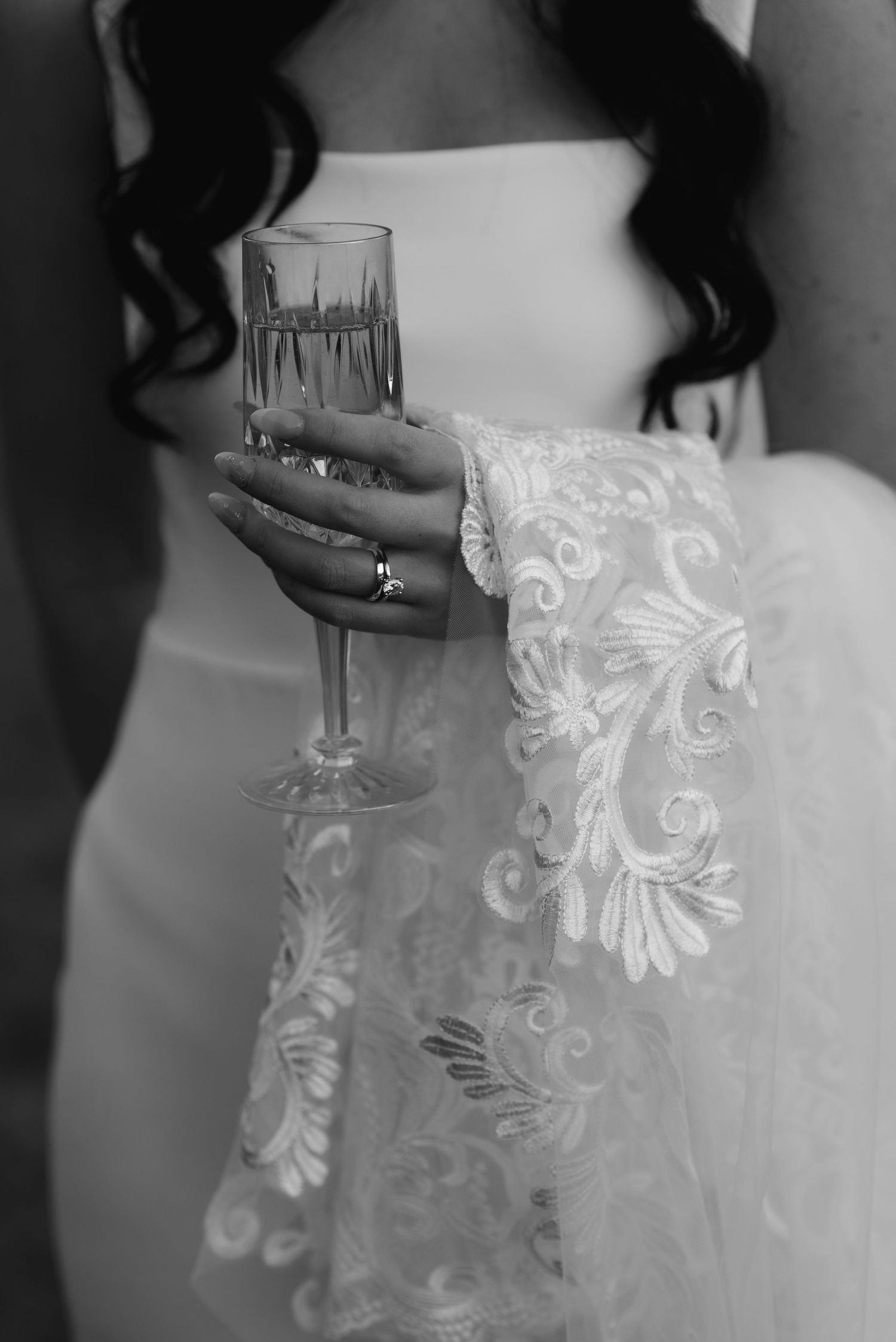Black and white photo of a bride in lace wedding dress holding a champagne flute with a diamond ring on her finger.