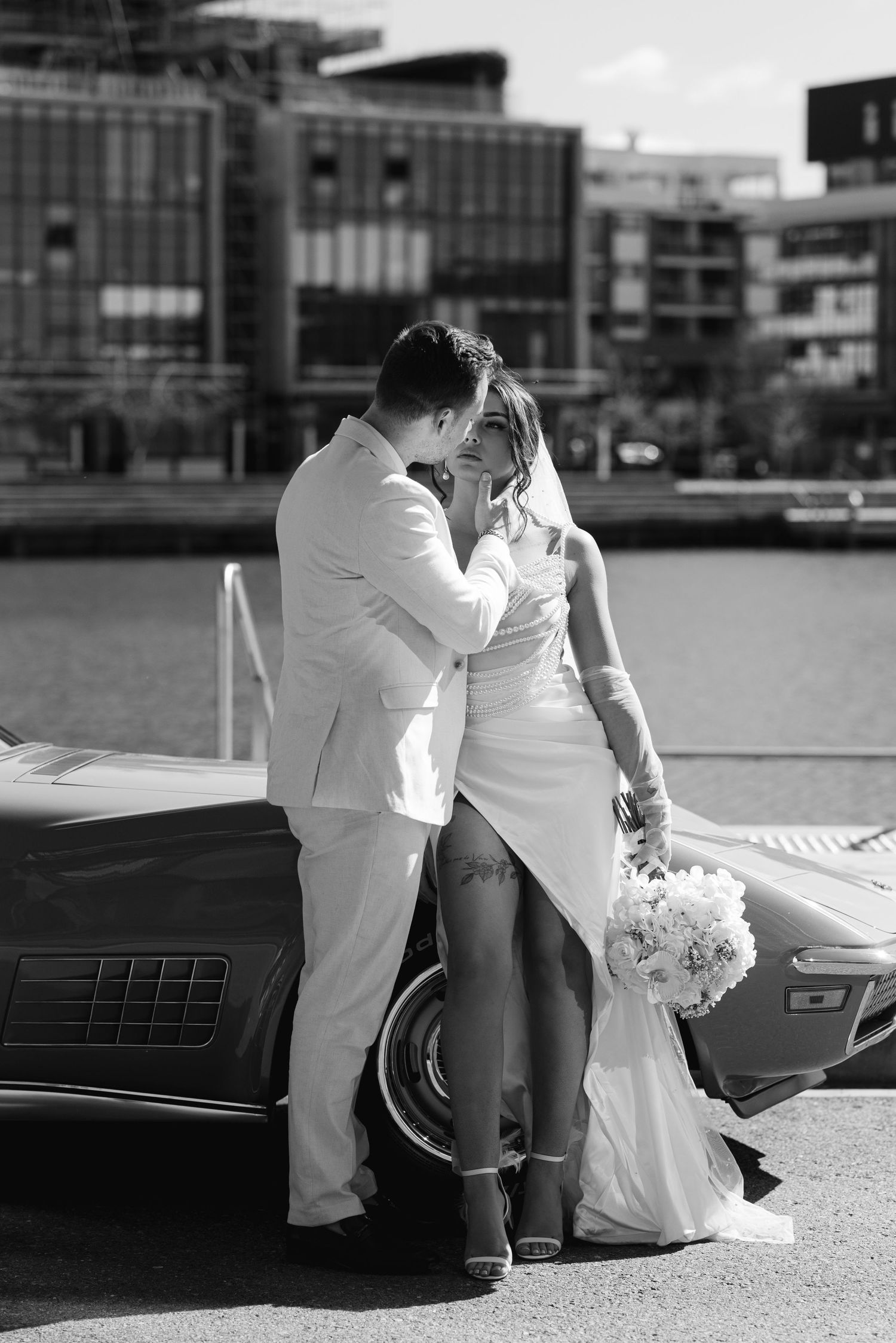 Black and white photo of a bride and groom kissing beside a classic car near a waterfront on their wedding day.