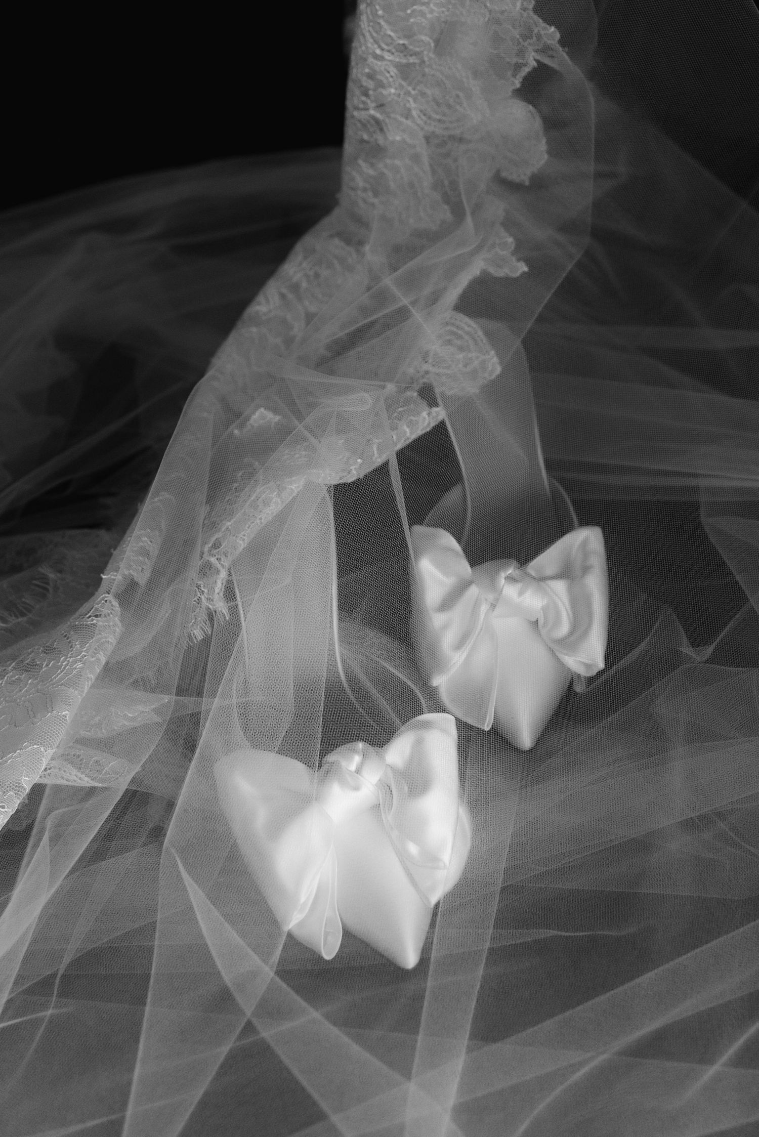 Black and white photo of elegant bridal satin heels with bows resting on a flowing wedding veil.