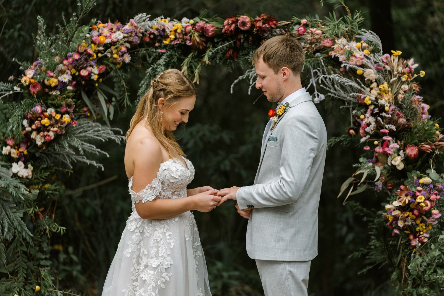 Bride and groom exchange rings under a floral arch adorned with colorful blooms during an outdoor wedding ceremony.