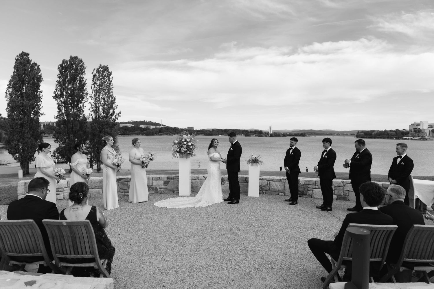A black and white panoramic view of guests seated at an outdoor wedding ceremony with tall trees in the background.