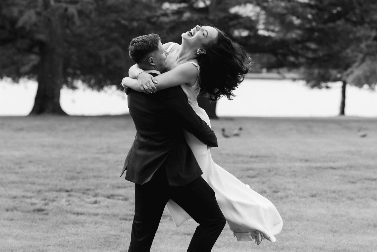 A romantic couple in formal wedding attire dances together in a park setting.