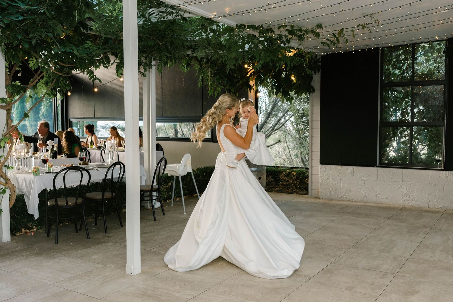 Bride in elegant white gown shares first dance at romantic indoor wedding reception with lush greenery decor.