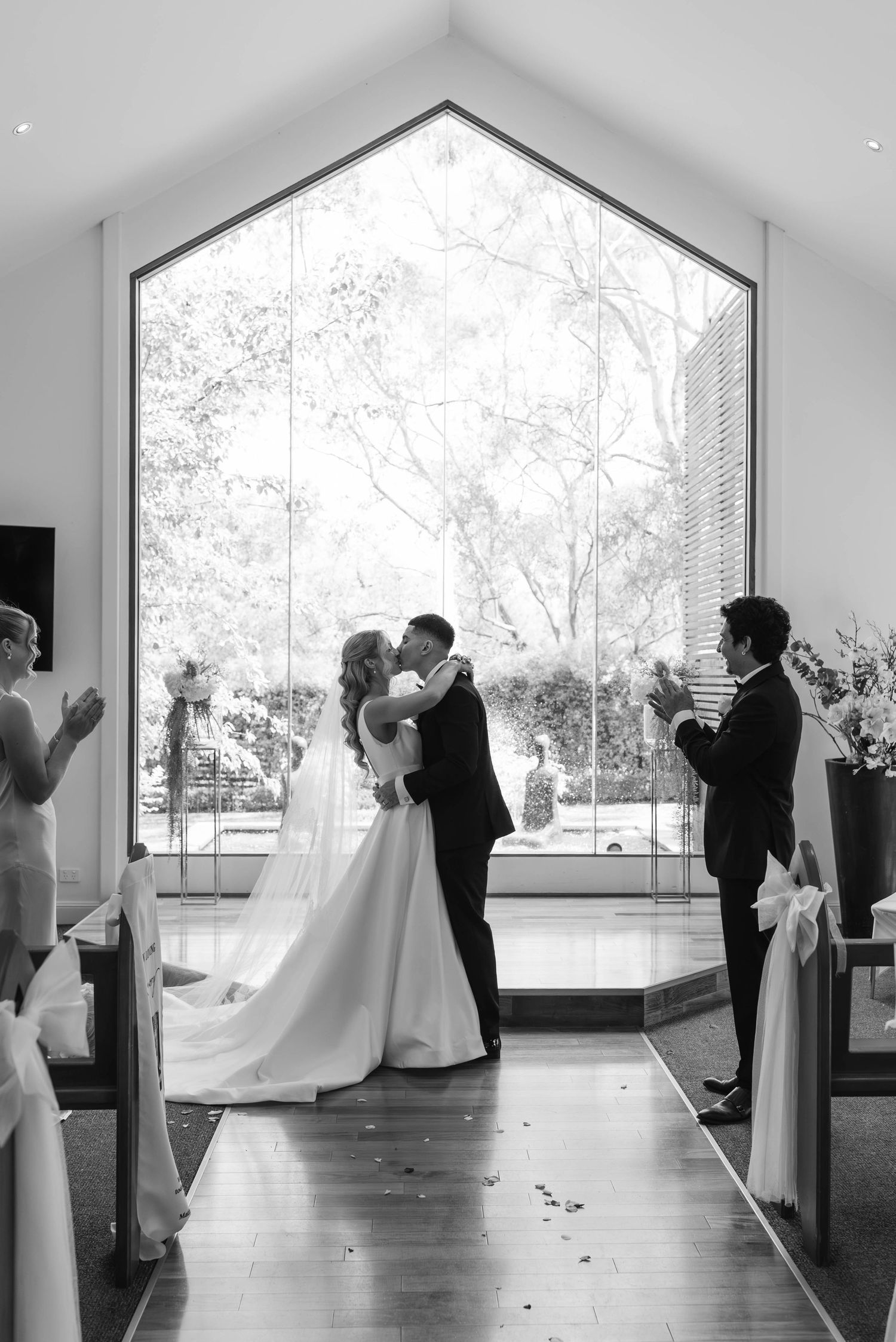 Bride and groom share first kiss at altar in elegant chapel with large arched window in black and white.