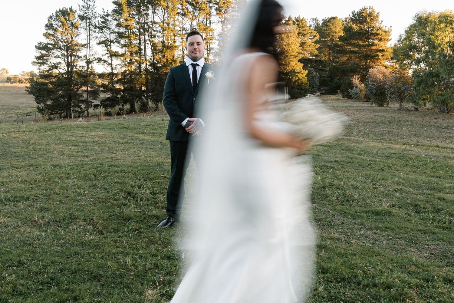 Groom in black suit stands in green field as blurred bride in white gown approaches holding bouquet at outdoor wedding.