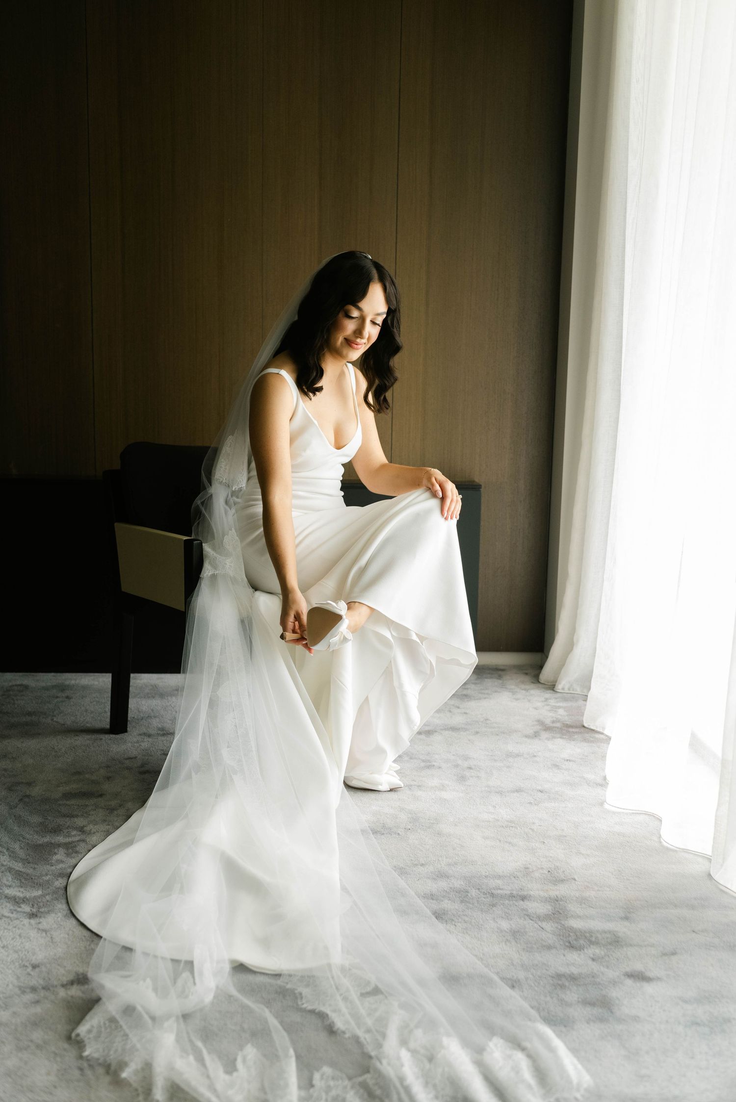 A bride in a flowing white dress sits elegantly by a window, with soft natural light illuminating the scene.