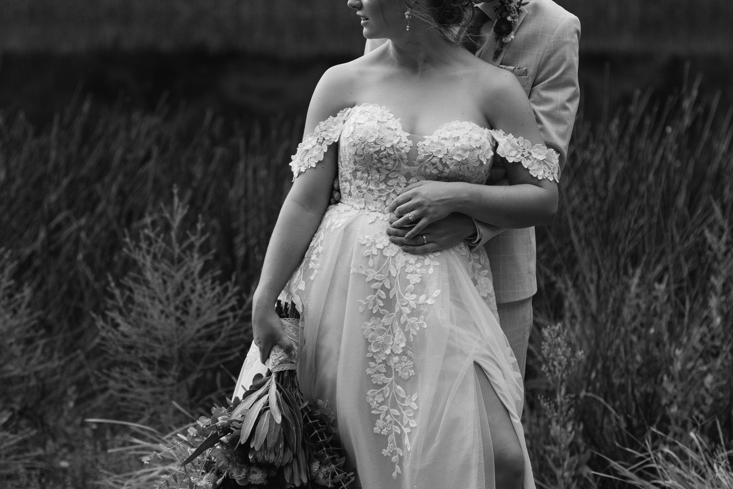 Black and white photo of a bride in lace off-shoulder gown holding bouquet, embraced by groom in field.