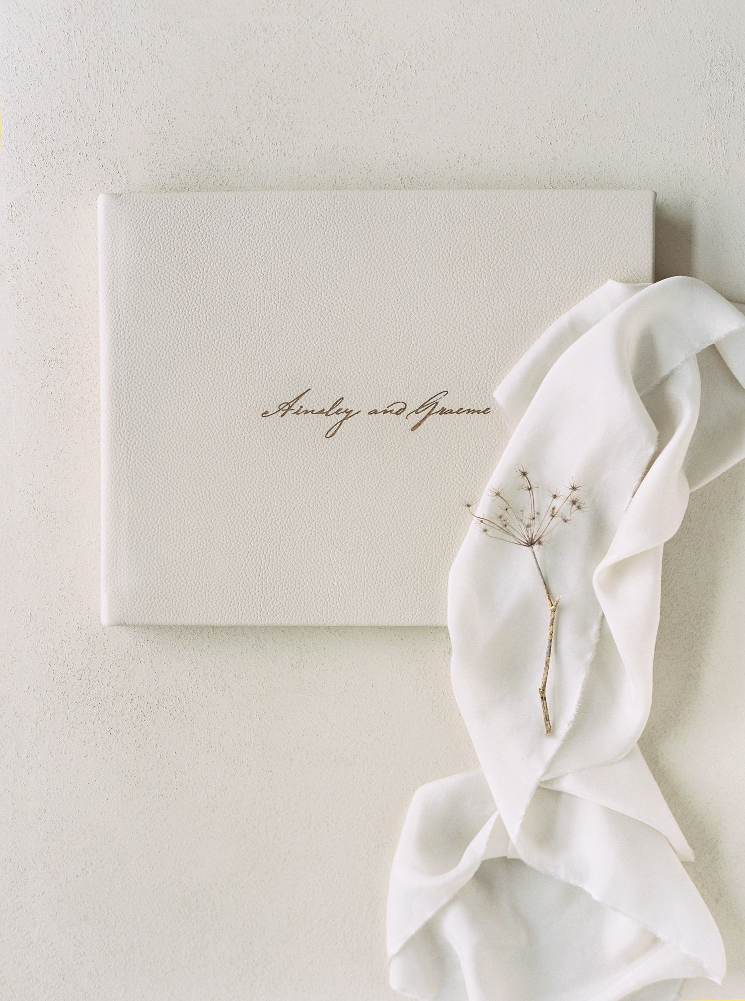 Elegant white linen wedding guest book with gold script personalization, styled with flowing silk ribbon and dried botanicals.