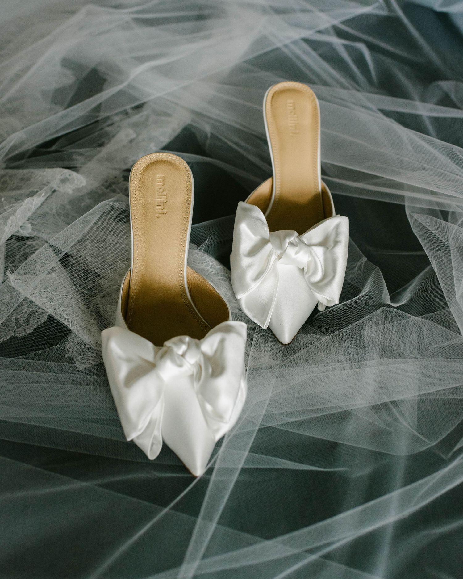 White satin bridal shoes with elegant bows rest on delicate tulle fabric in a dreamy composition.