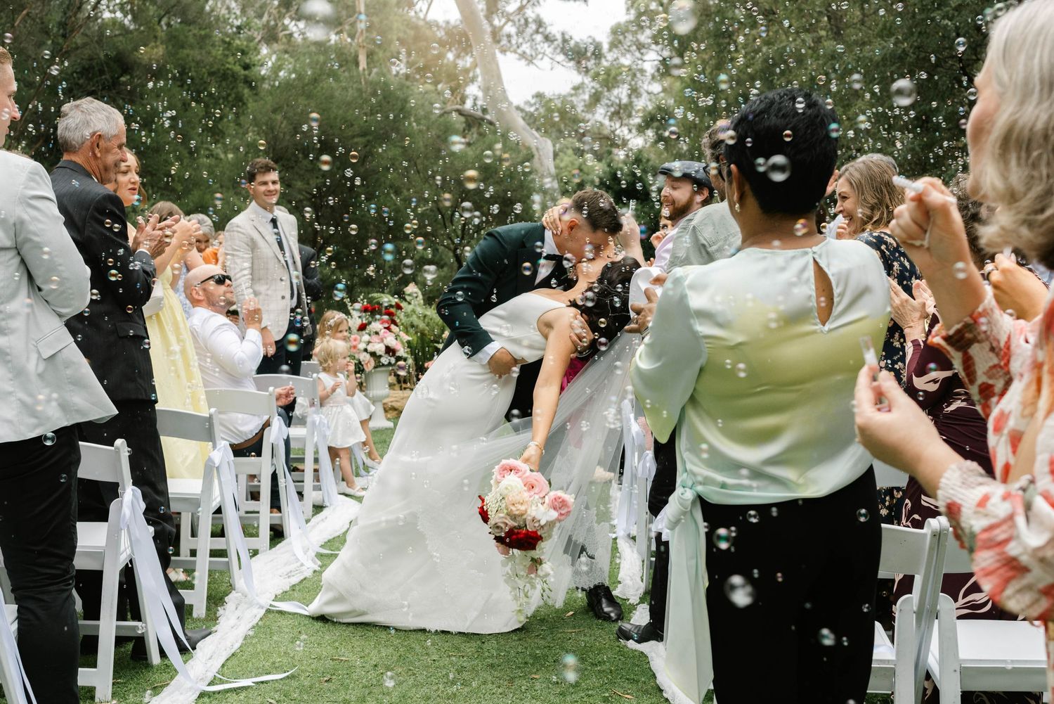 Wedding guests celebrate by blowing bubbles during an outdoor garden ceremony with white chairs.