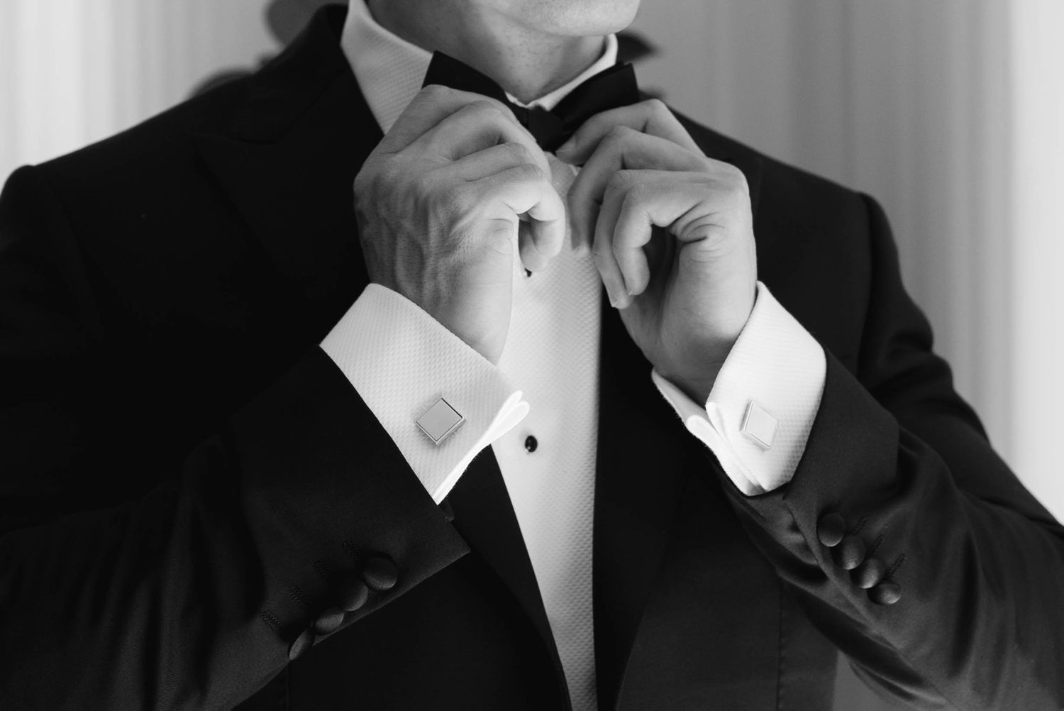 Black and white photo of a man in a tuxedo adjusting his bow tie, showcasing elegant formal attire.