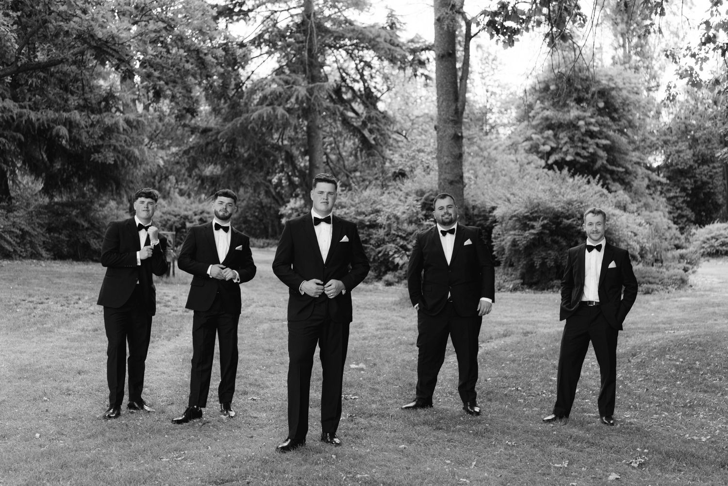 Wedding party members in black tuxedos walking in formation across a garden setting.