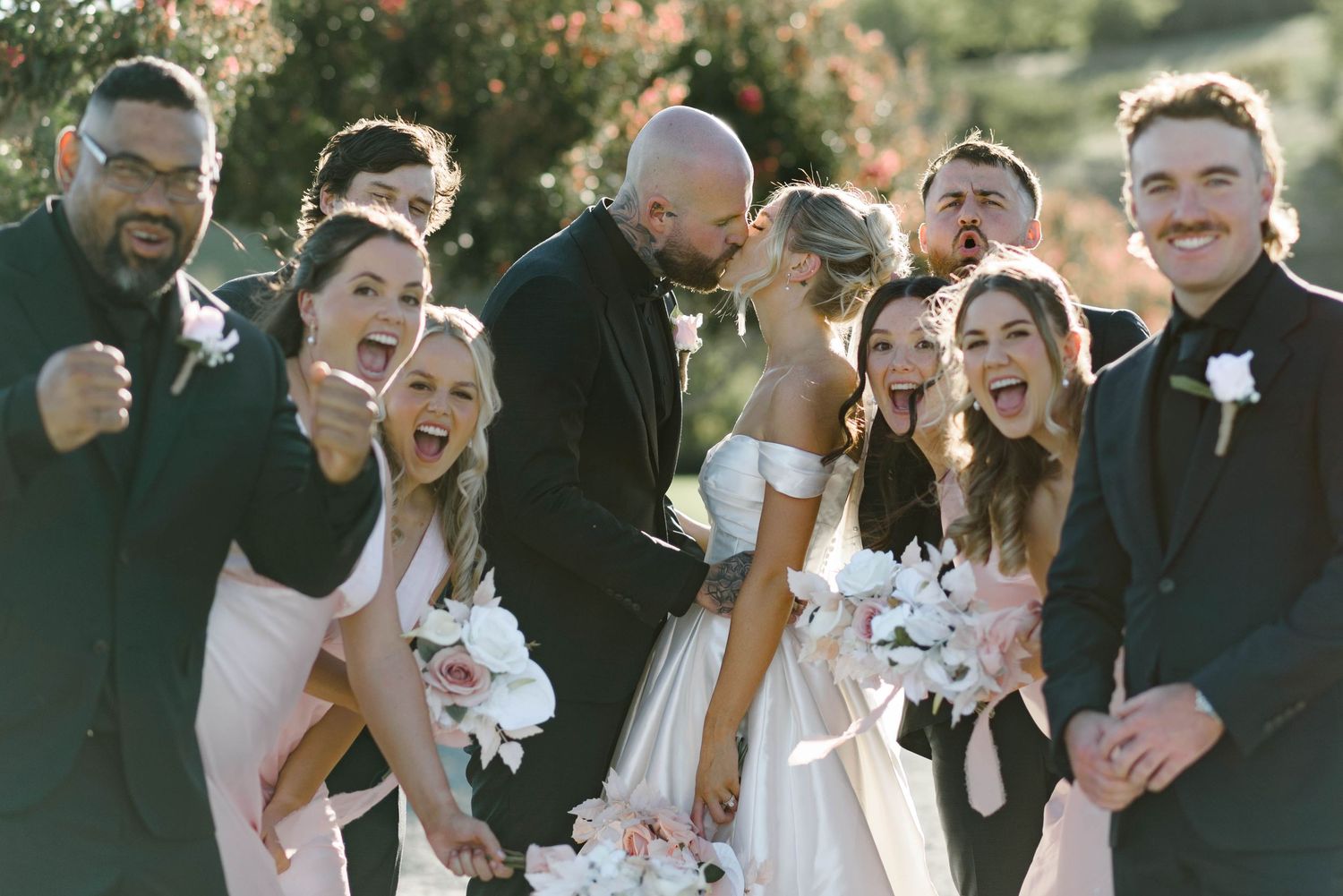 A joyful wedding party in formal attire laughs together during an outdoor celebration.