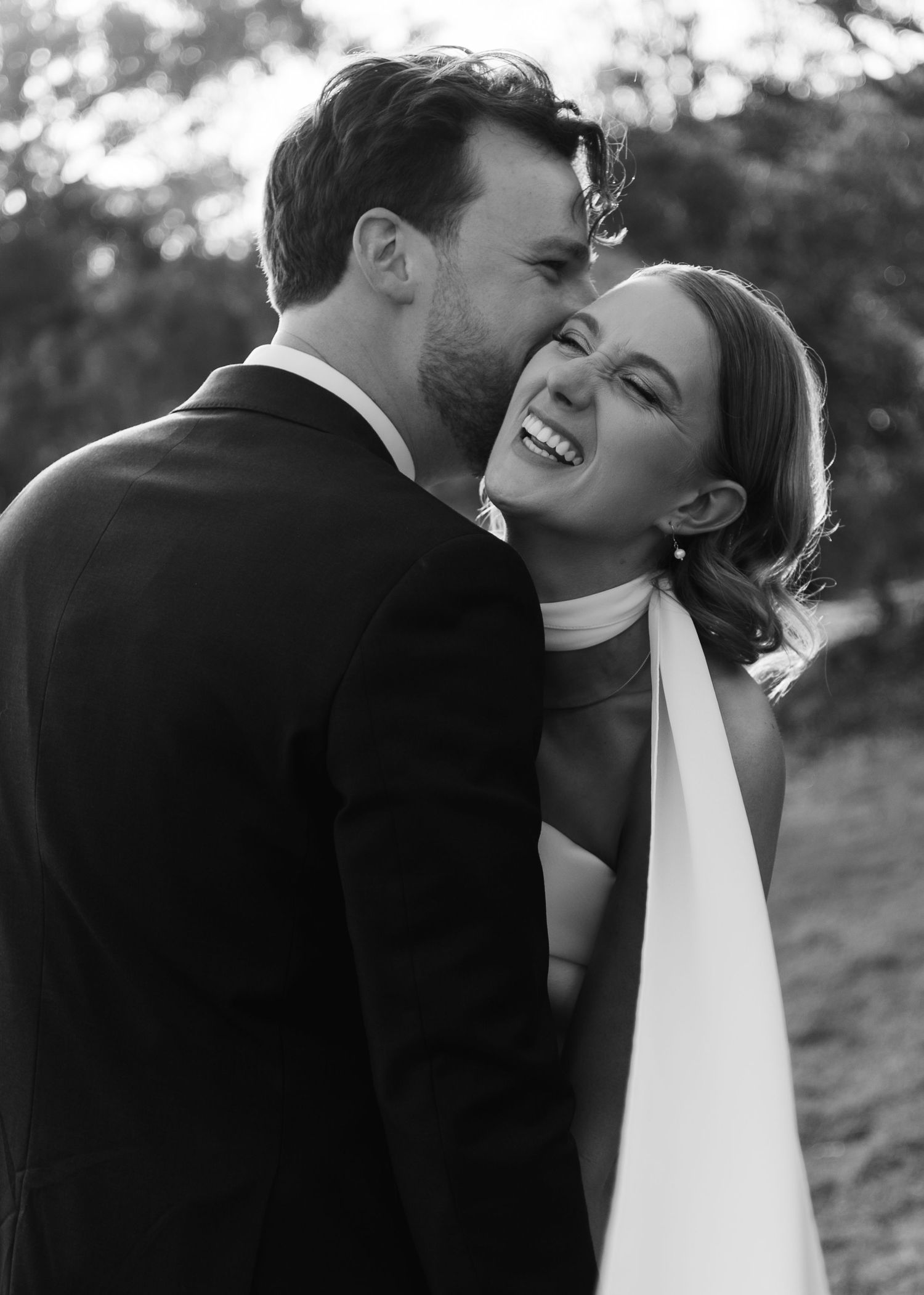 Black and white wedding photo of a laughing bride in white dress as groom whispers in her ear outdoors.