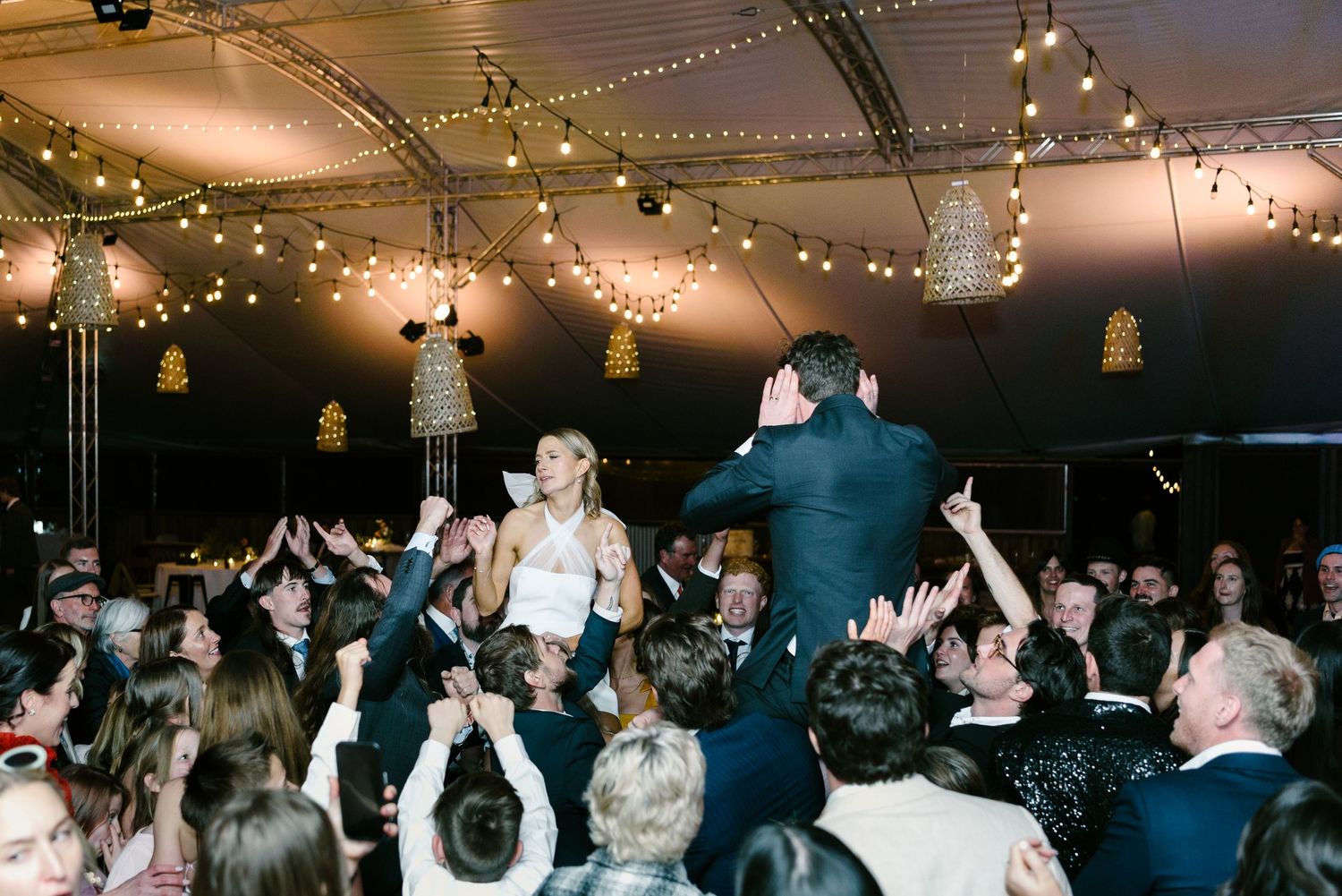 Bride and groom lifted by guests during lively wedding reception under string lights in elegant tent venue.