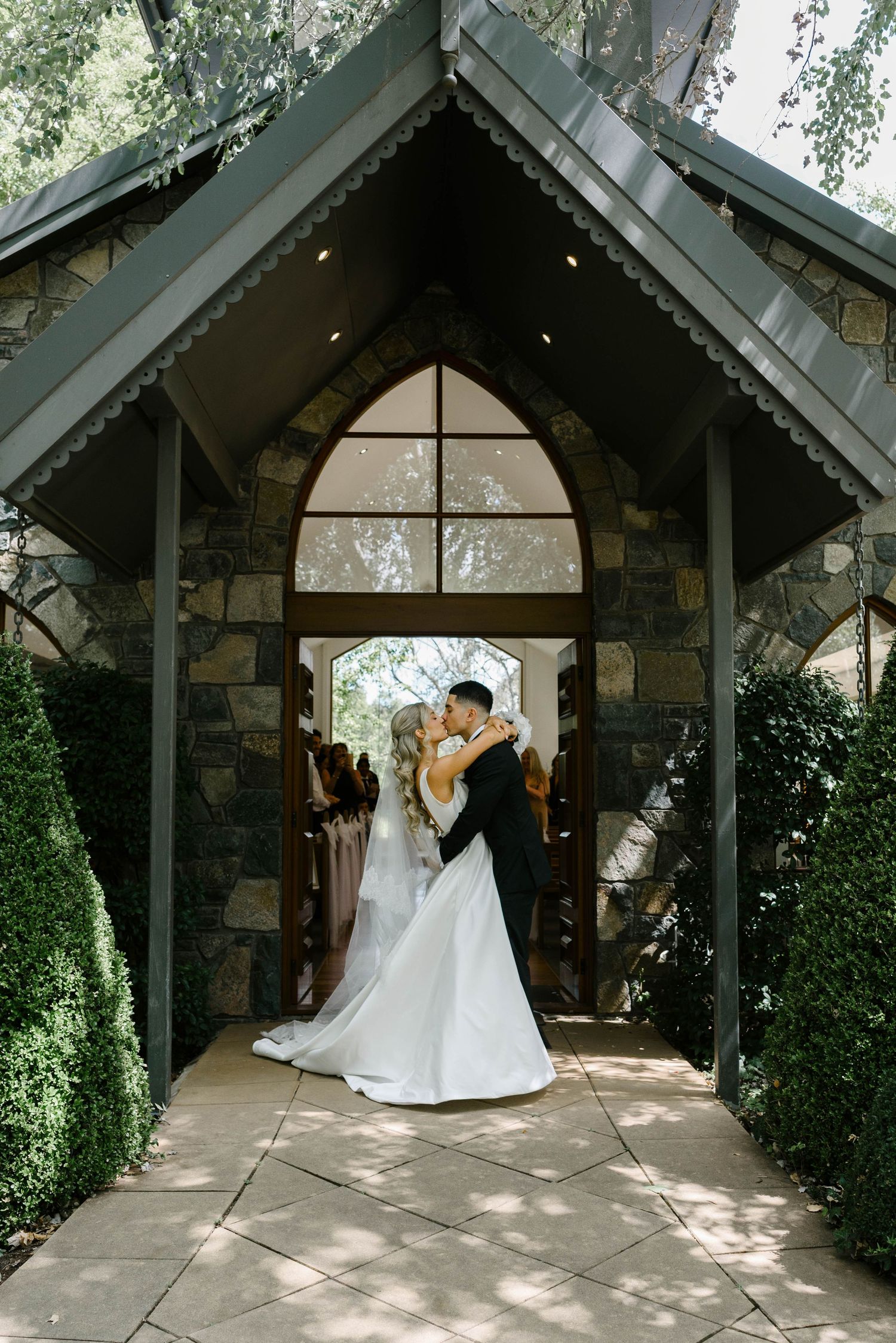 Newlyweds share first kiss at entrance of stone chapel surrounded by lush greenery.