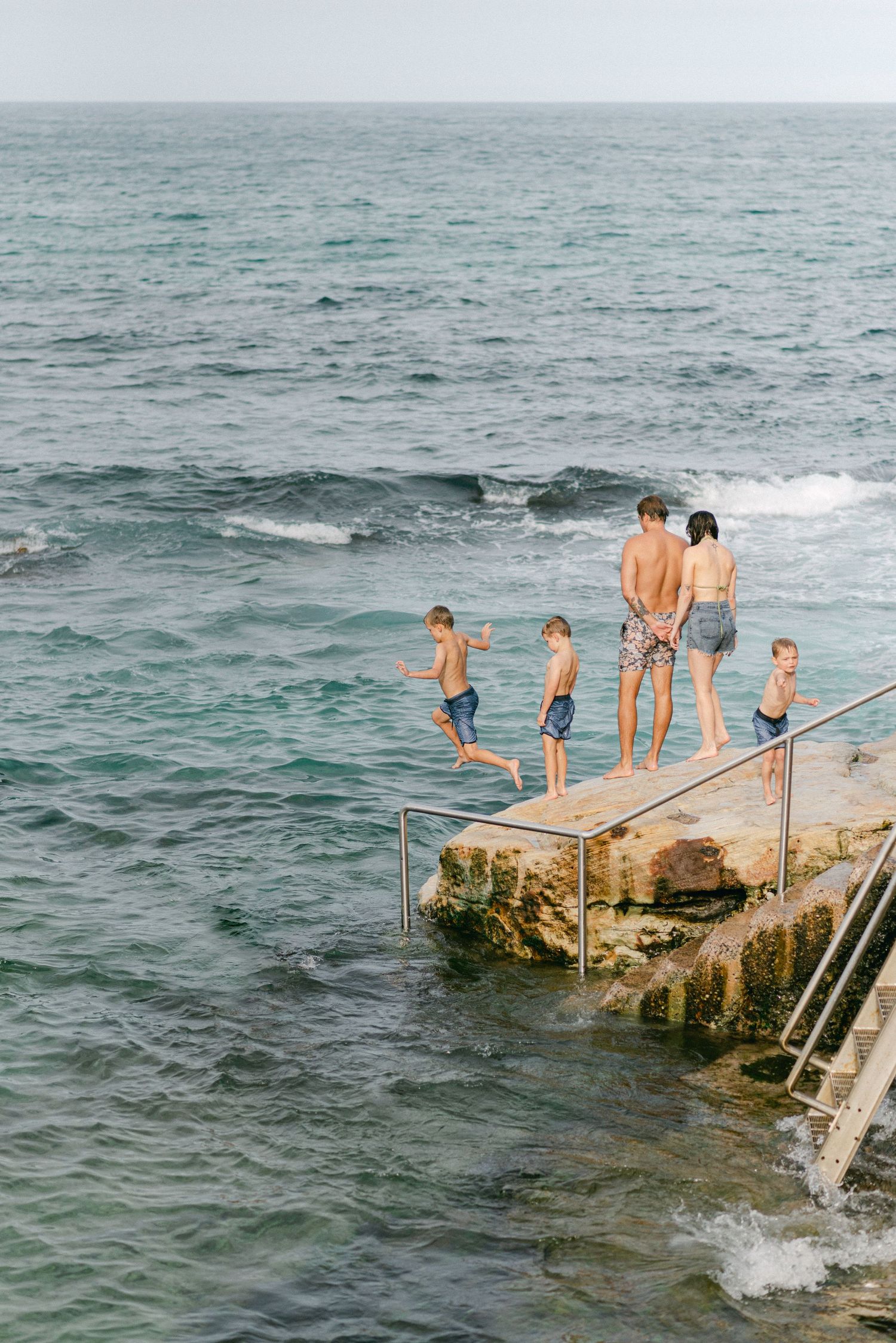 People jumping off a concrete pier into the ocean on a sunny day.