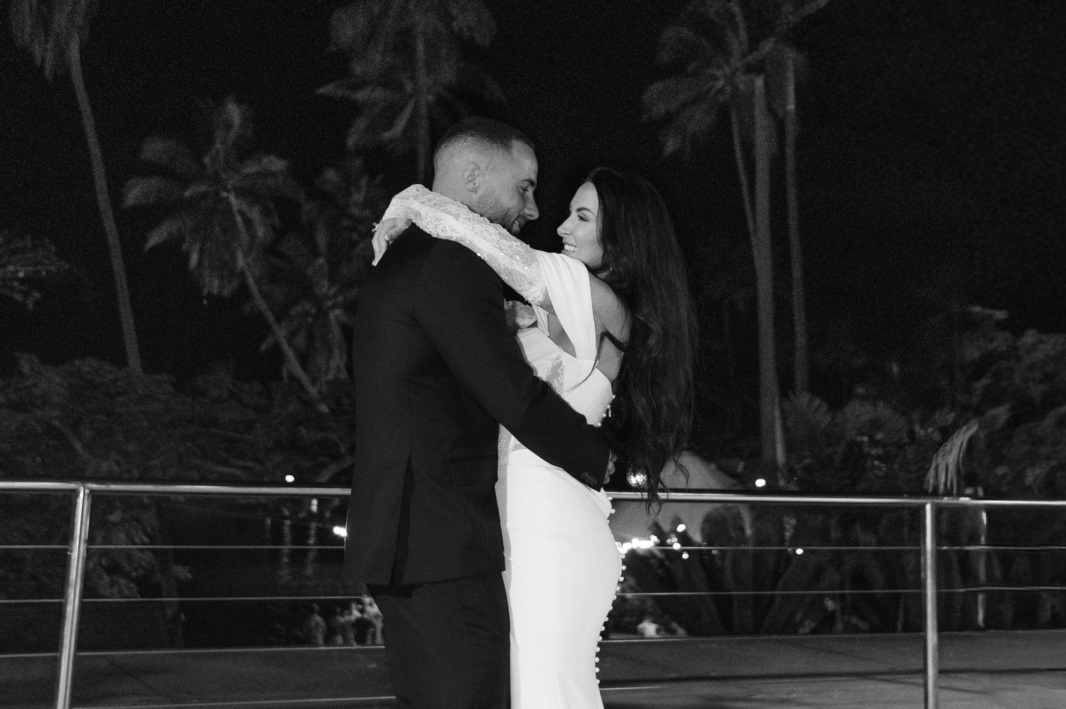A couple embraces romantically on a balcony at night, surrounded by palm trees, in a elegant black and white photo.