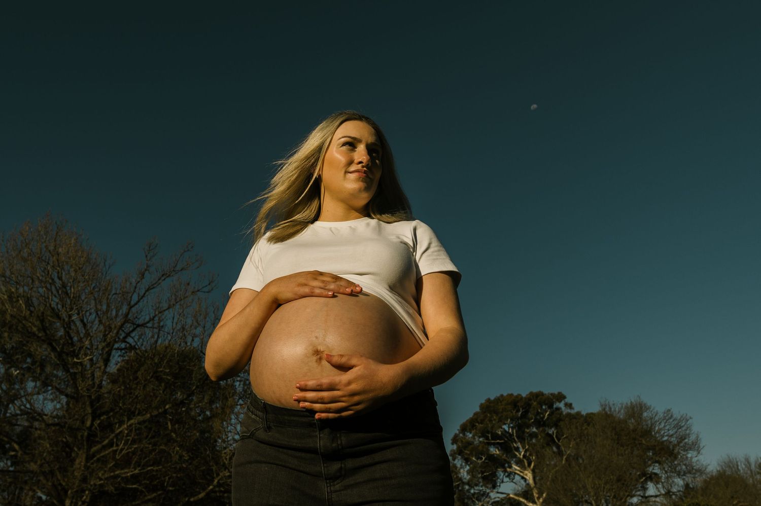 Pregnant woman in white crop top cradling baby bump outdoors at dusk with trees and blue sky.
