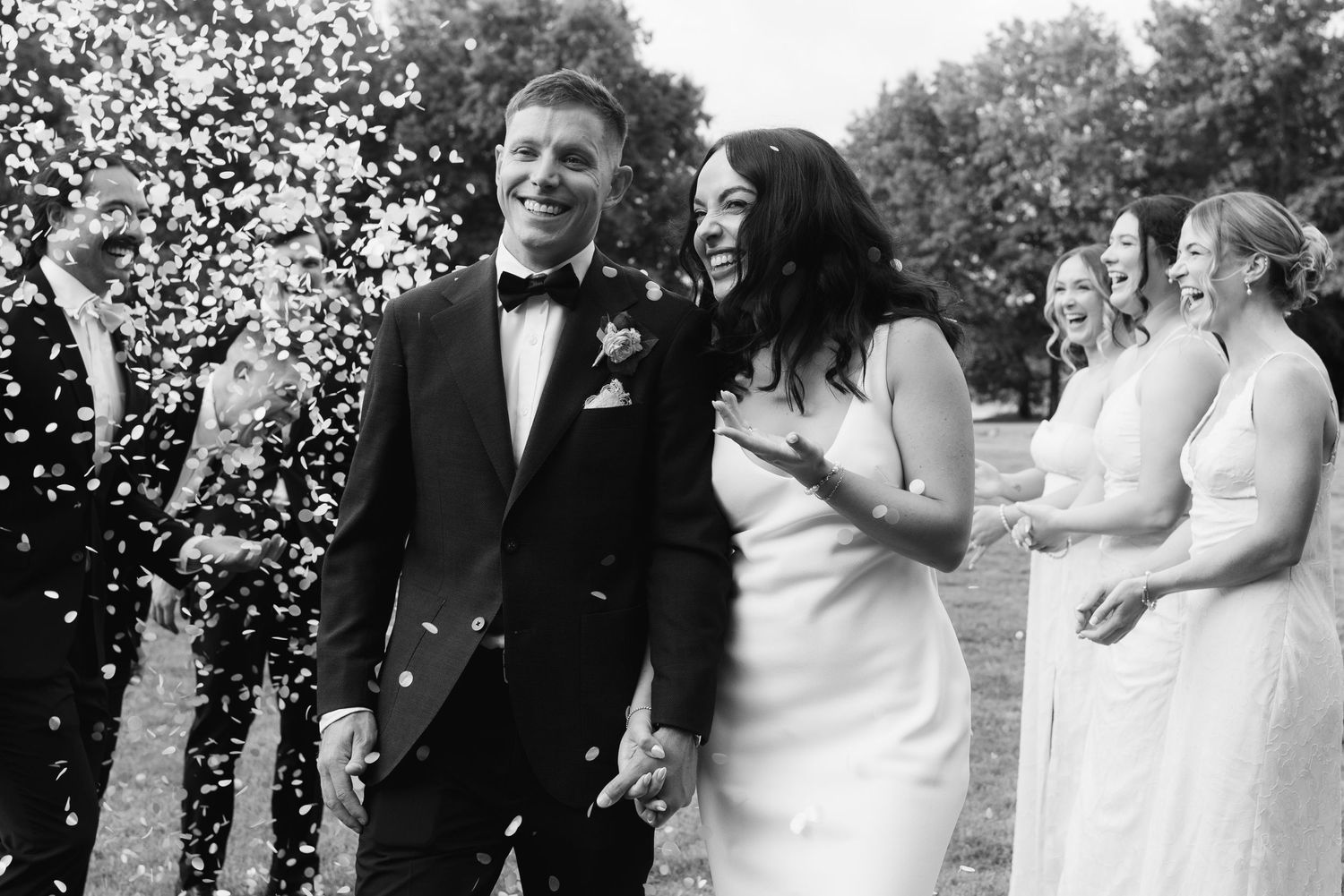 Black and white photo of a joyful bride and groom walking through confetti shower surrounded by wedding guests.