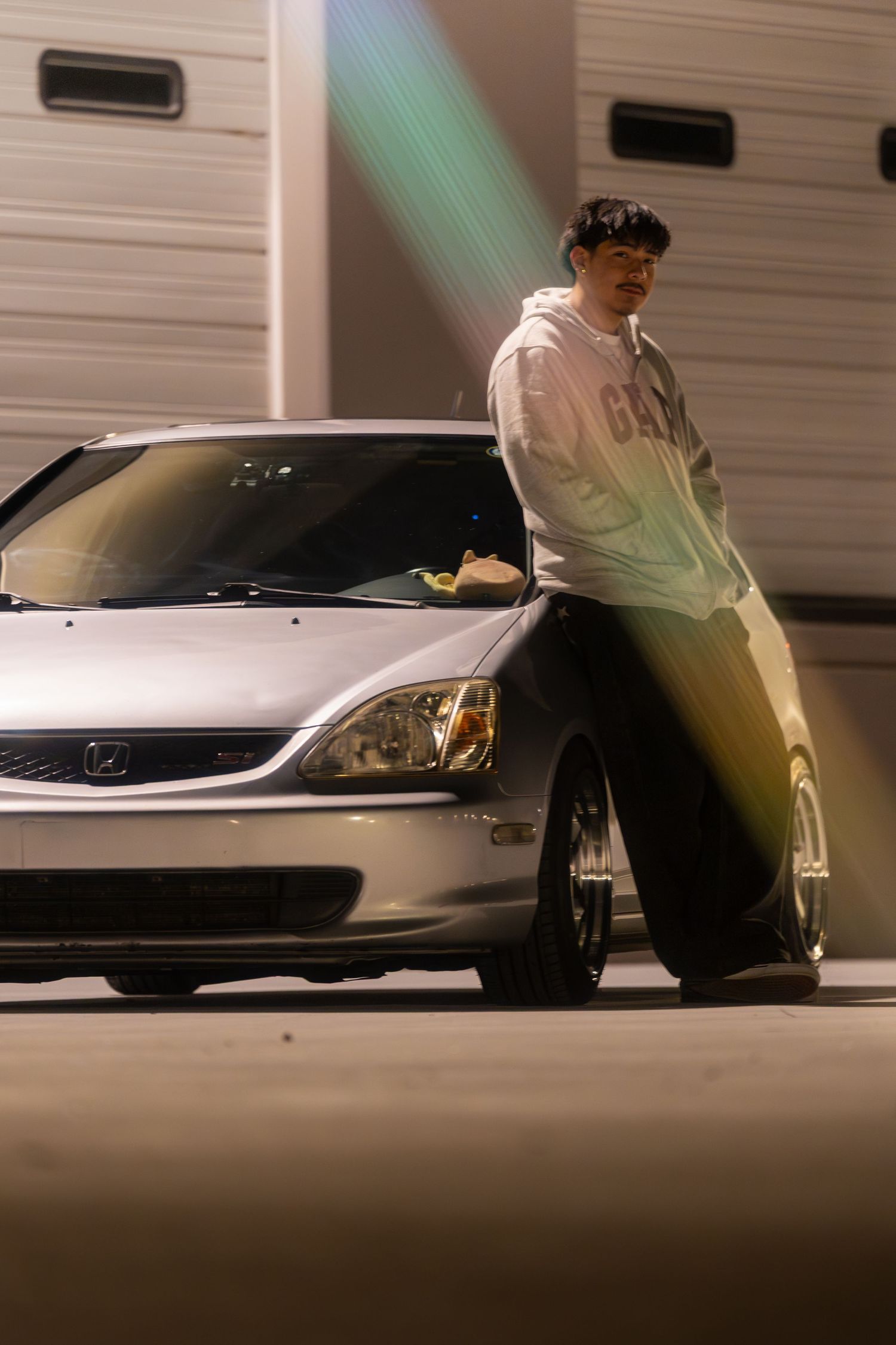 Young man in white hoodie leaning against a silver Honda Civic at night with dramatic light rays in background.