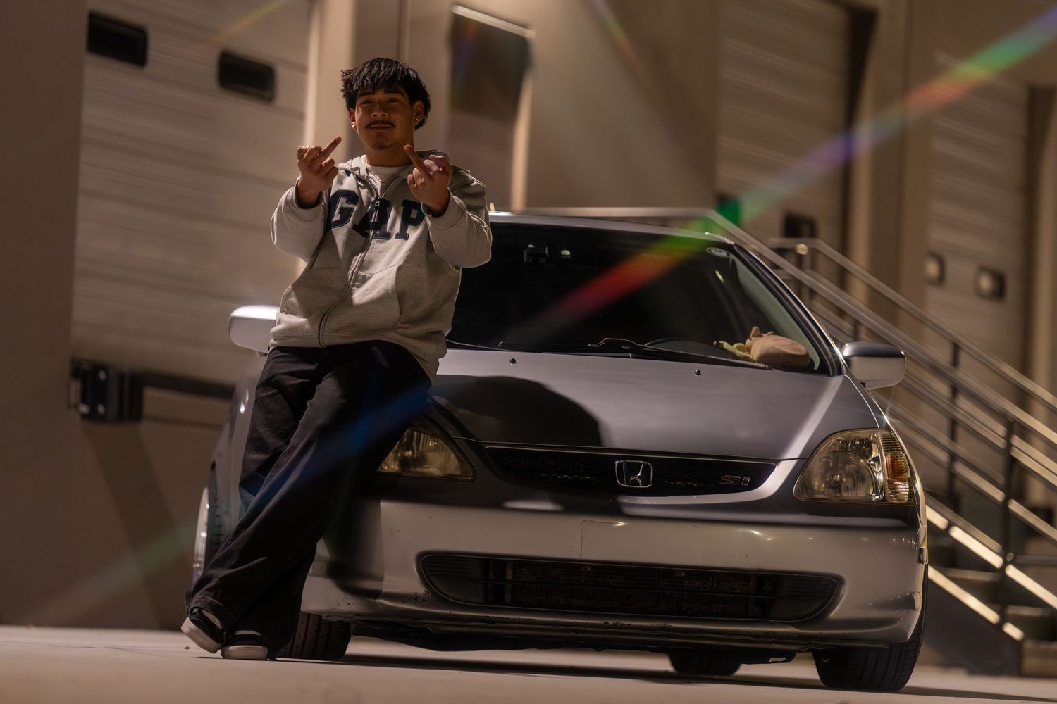 Young man posing on silver Honda Civic at night with colorful lens flare effect in parking garage.