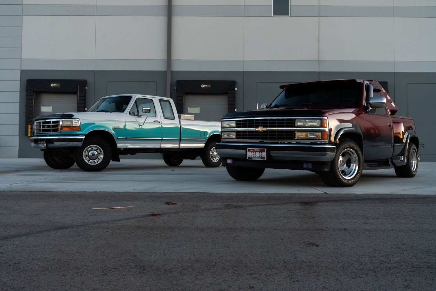 Row of classic pickup trucks and cars parked against industrial building wall.