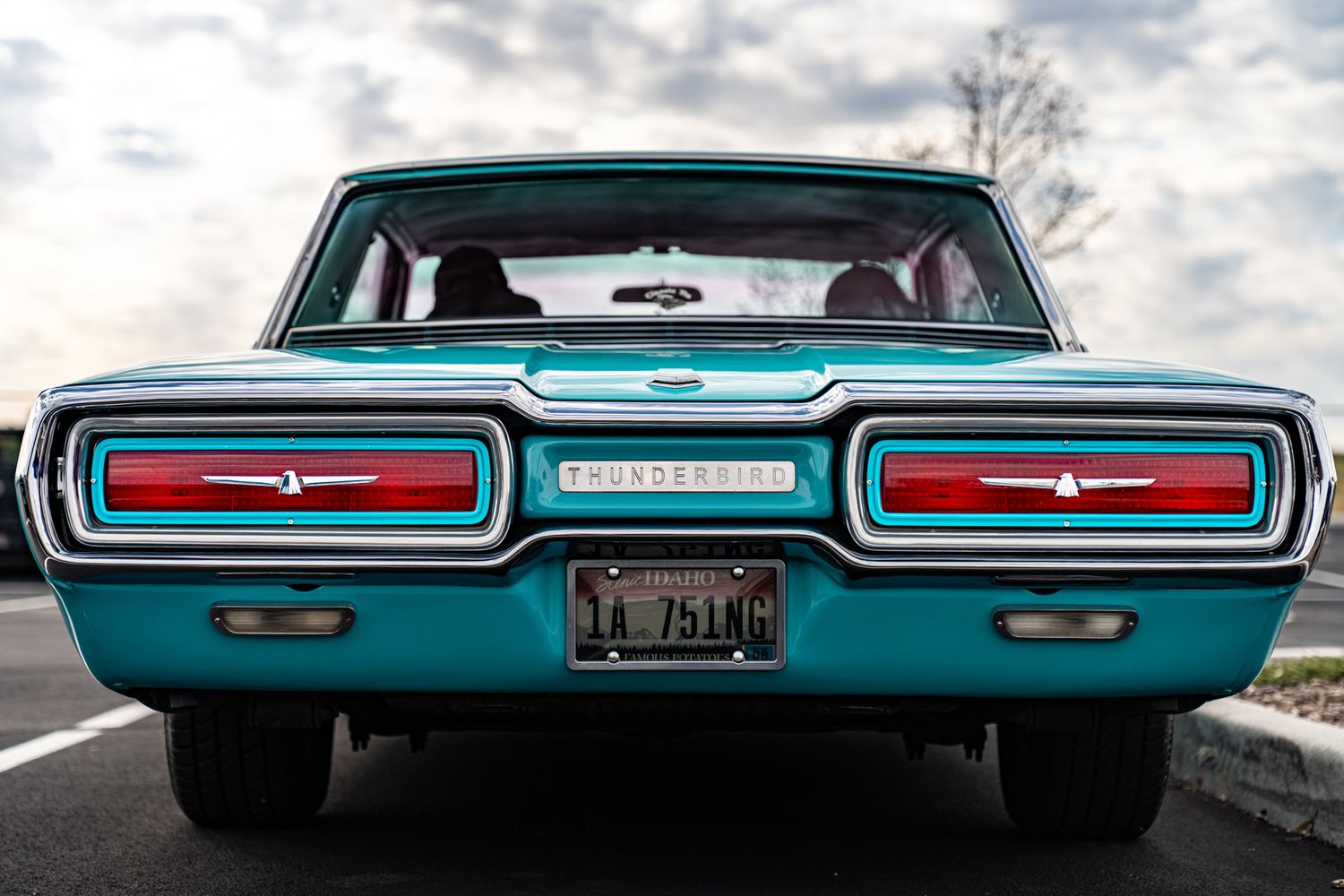 Rear view of a classic teal Ford Thunderbird muscle car with red tail lights parked on a road under cloudy skies.
