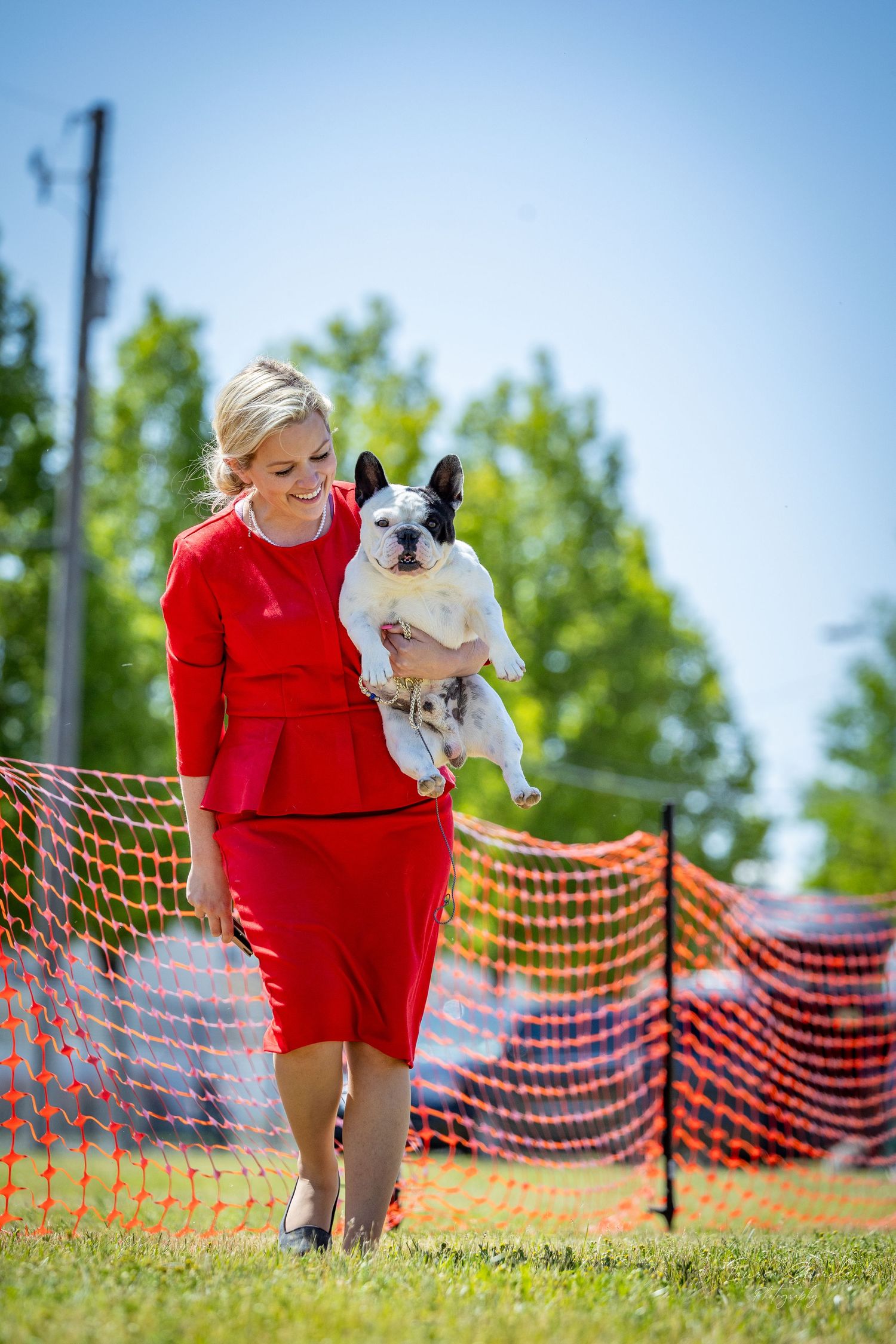 A small white dog runs alongside someone in red clothing on a sunny day with orange safety fencing visible.