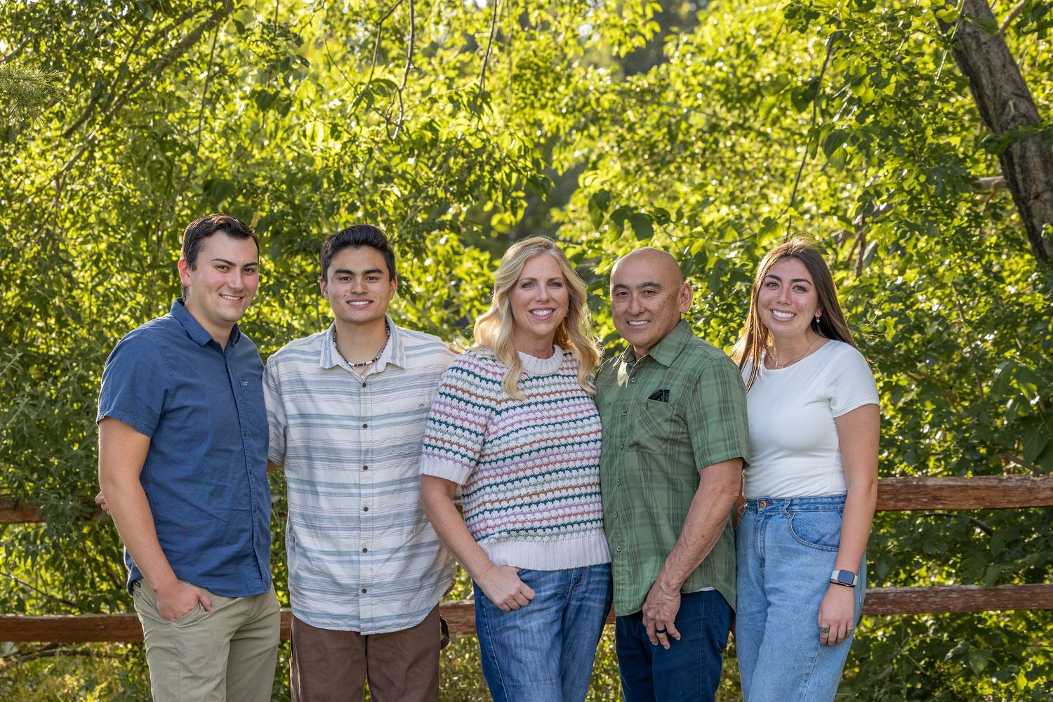 Family group poses together on wooden deck with lush green trees in background.