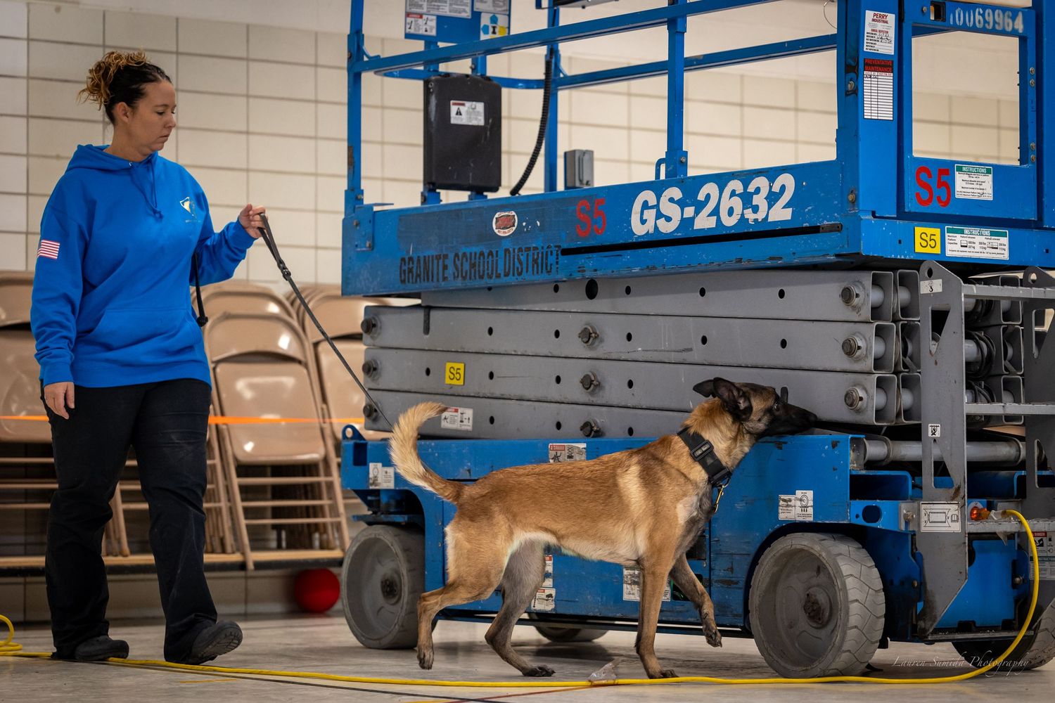 Dog handler works with canine partner near blue industrial lift equipment.