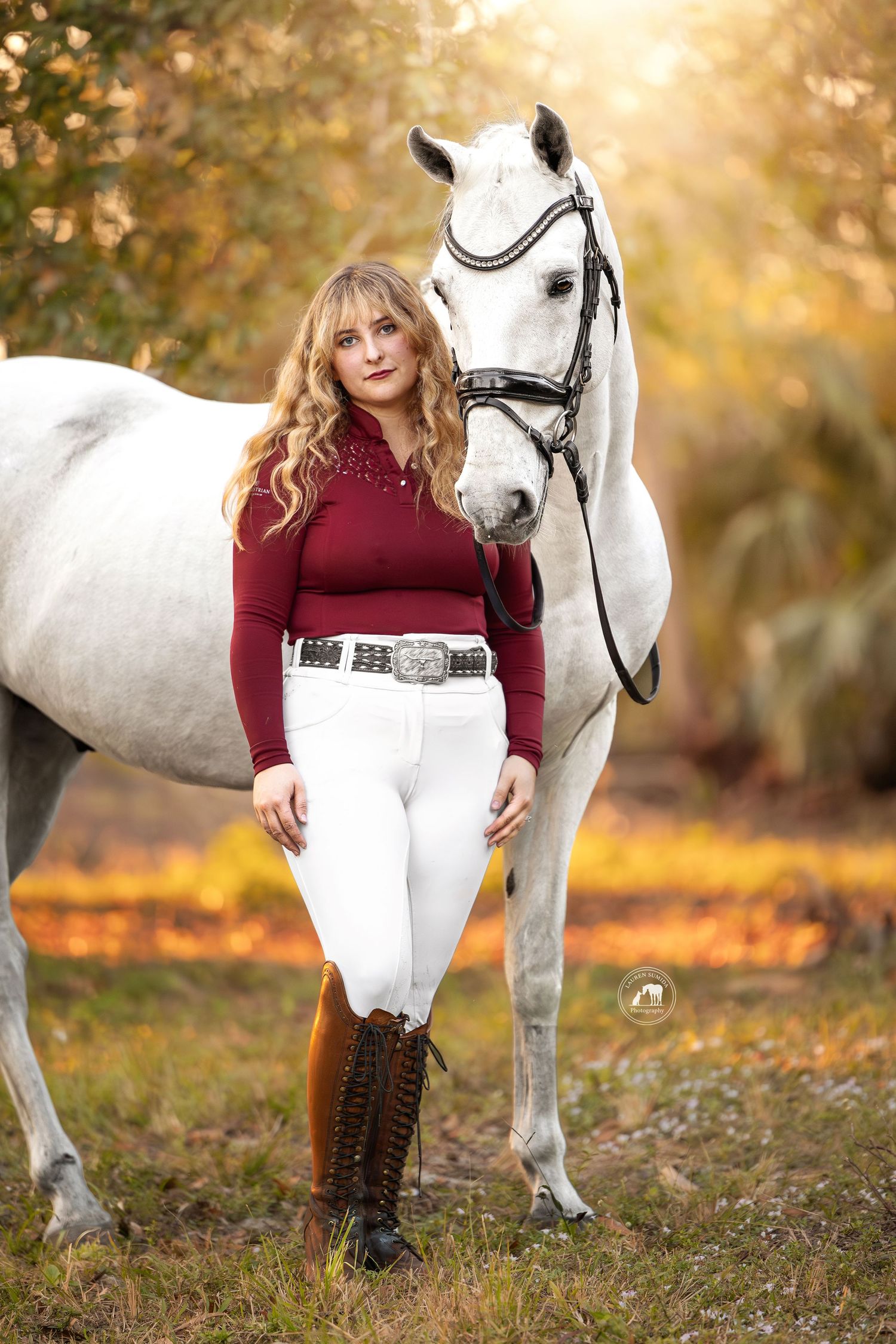 Woman in a burgundy top, white riding breeches and tall brown boots stands beside a bridled gray horse in a golden-lit autumn field.