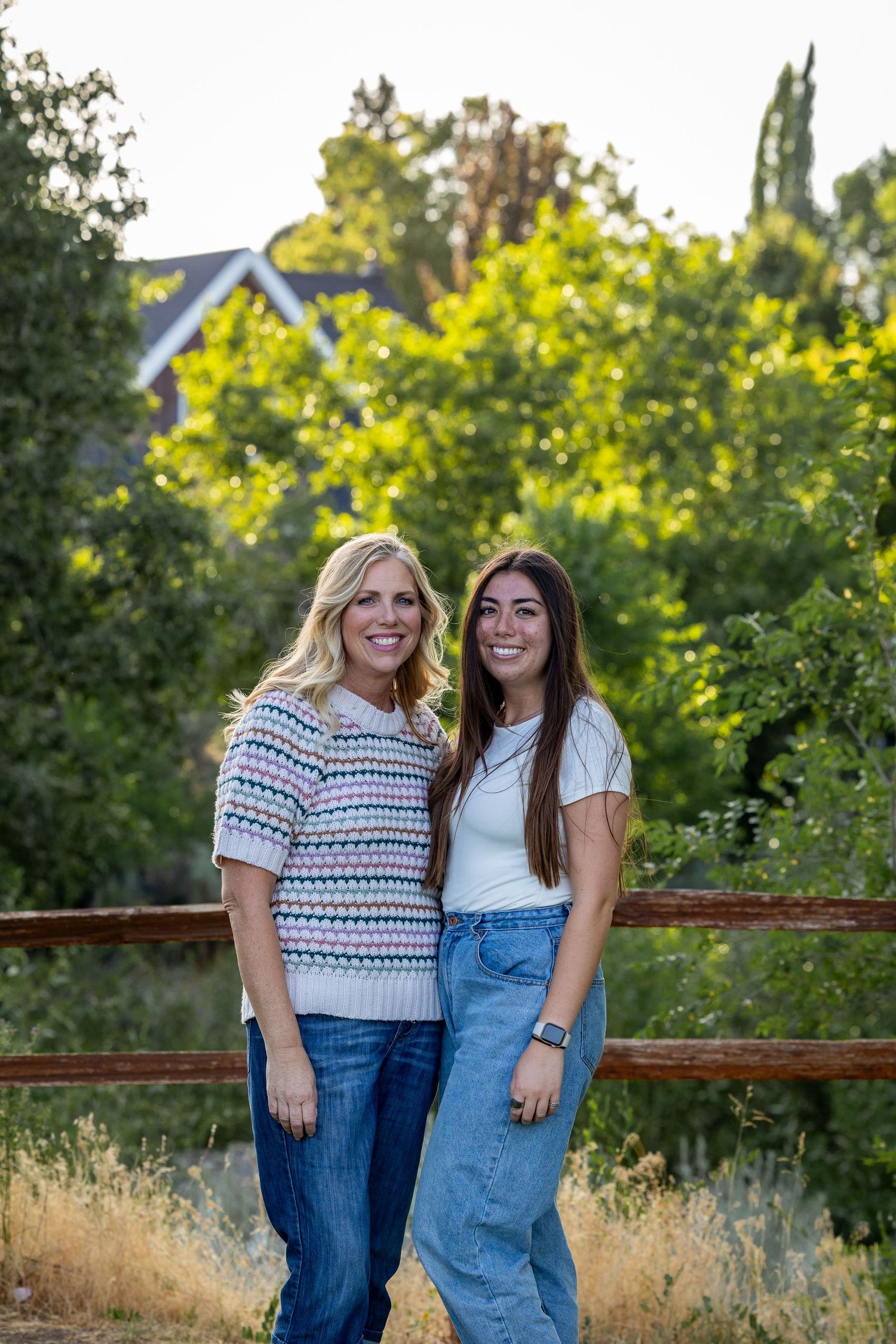 Two people pose together on wooden railing with green summer foliage in background.