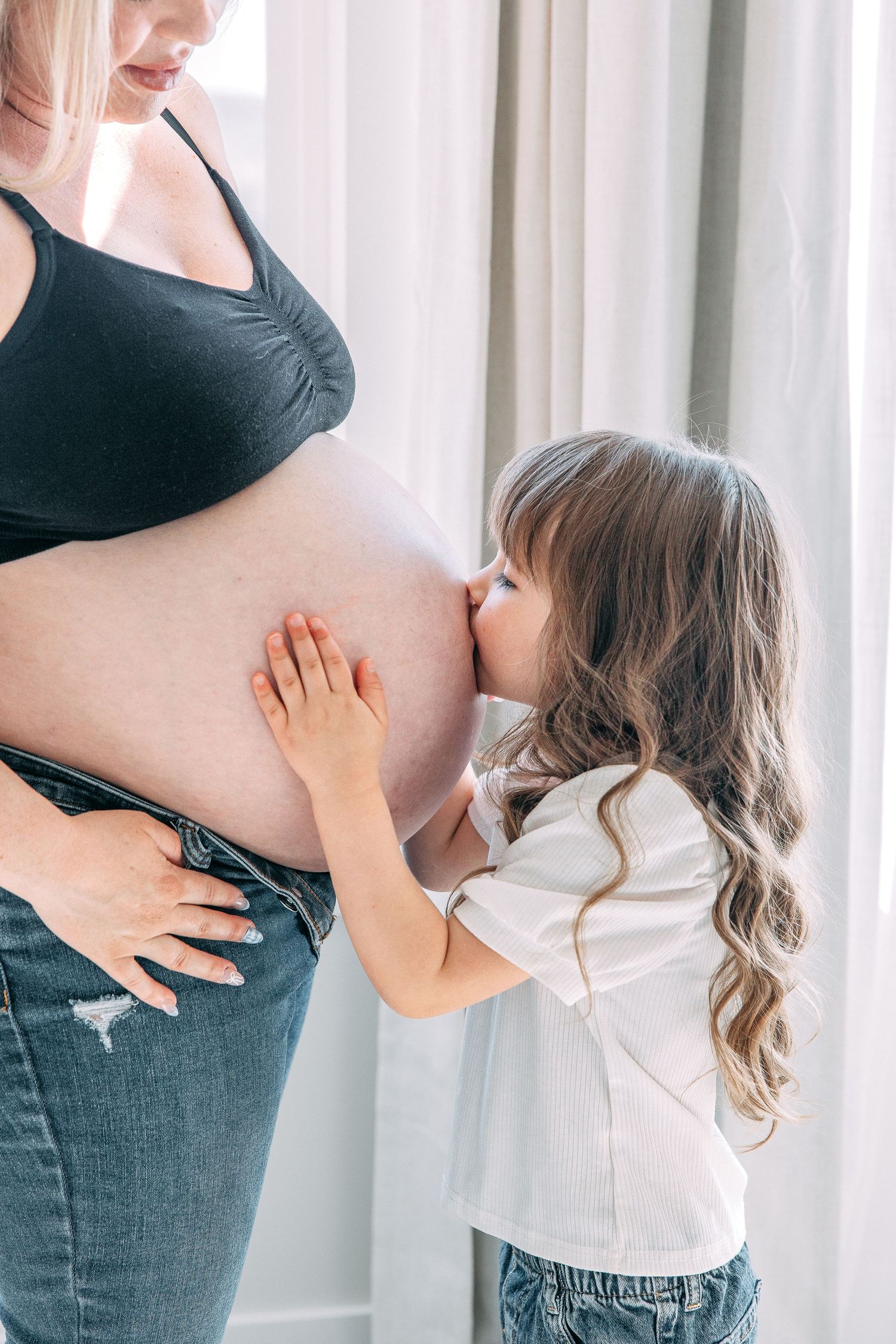 Child kissing pregnant belly in indoor maternity photo shoot.