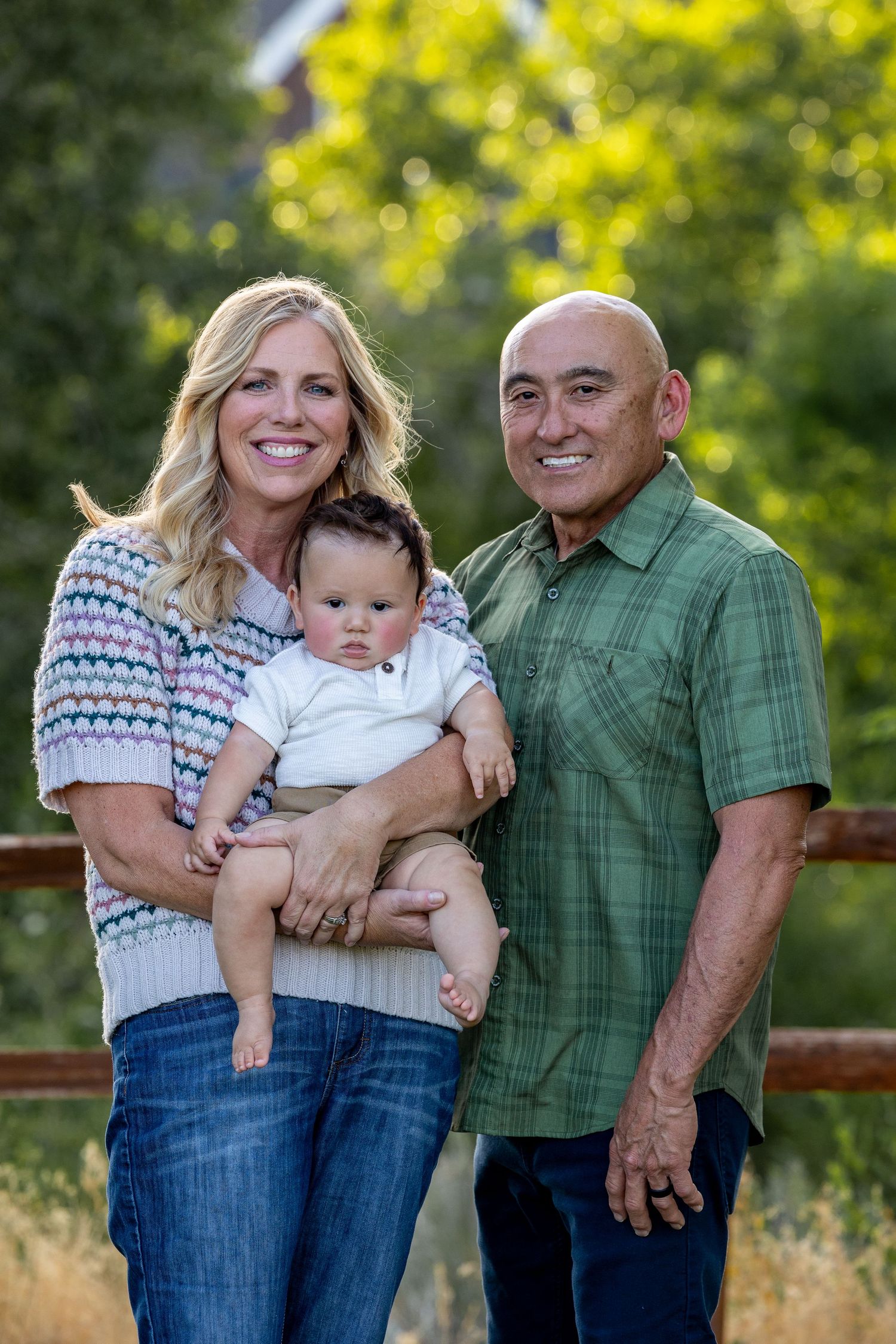 Family poses with baby on wooden deck during golden hour with trees in background.