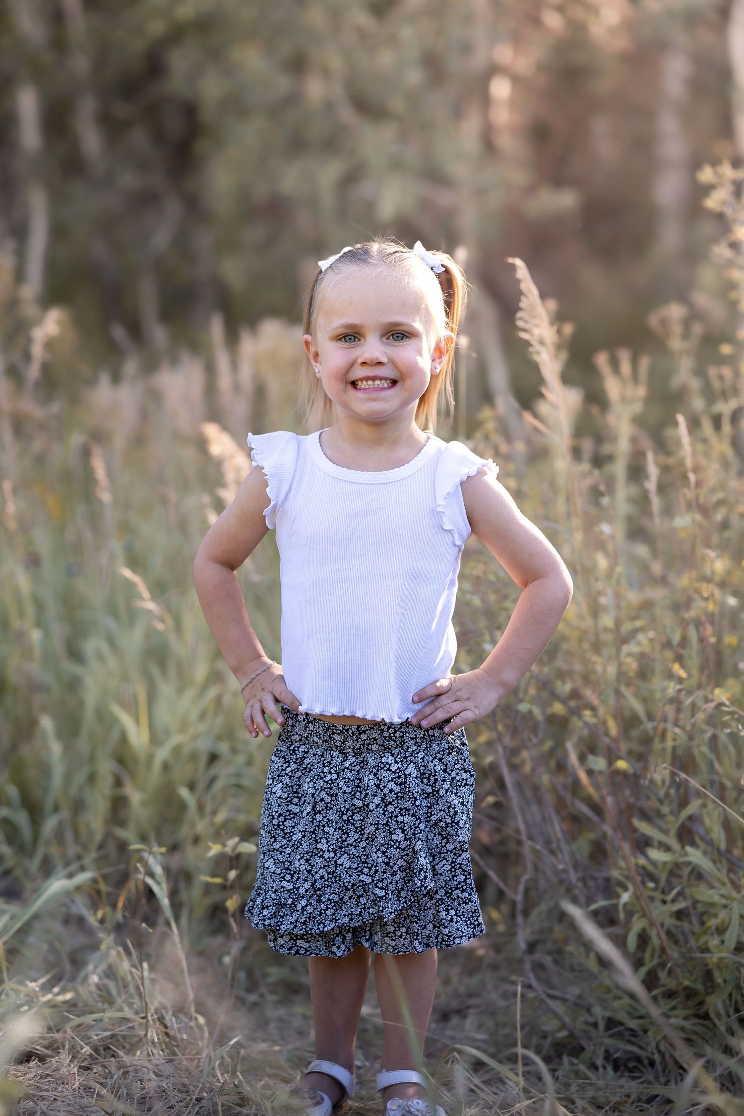 Young child in white top and patterned skirt poses playfully in meadow at golden hour.