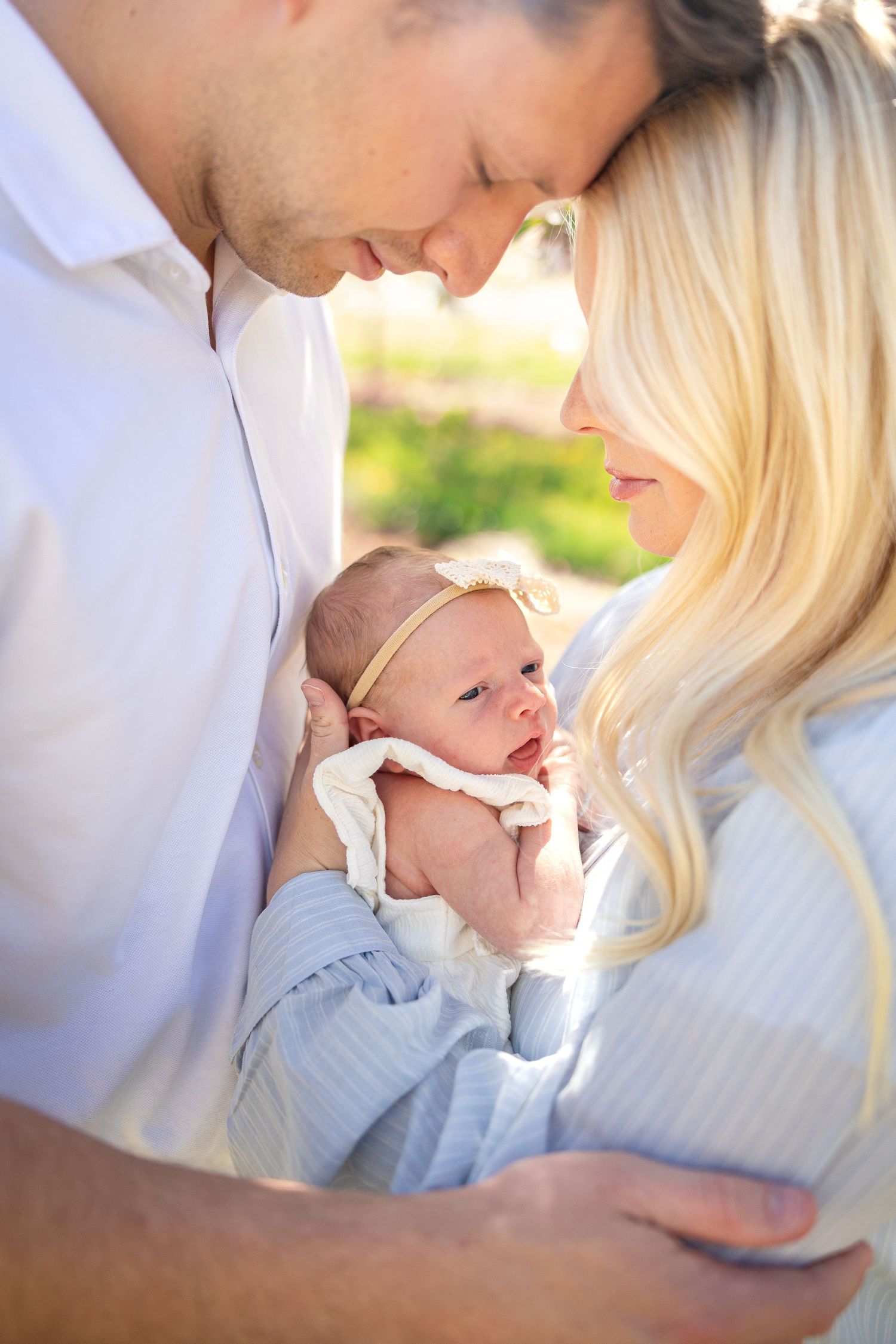 Parents cradling newborn baby outdoors in spring setting with soft lighting.