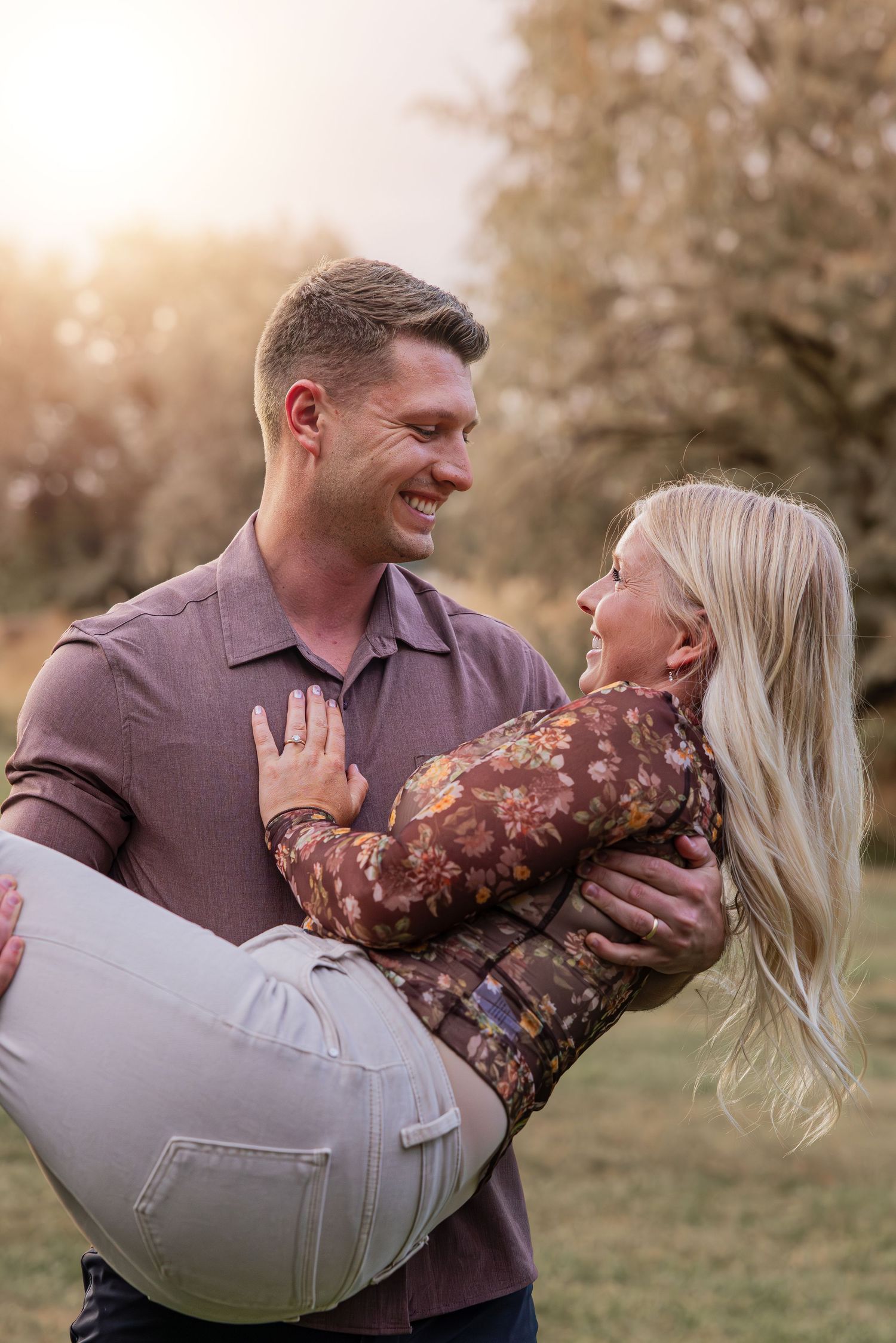 Playful couple embracing and laughing while posing in a grassy field at sunset.
