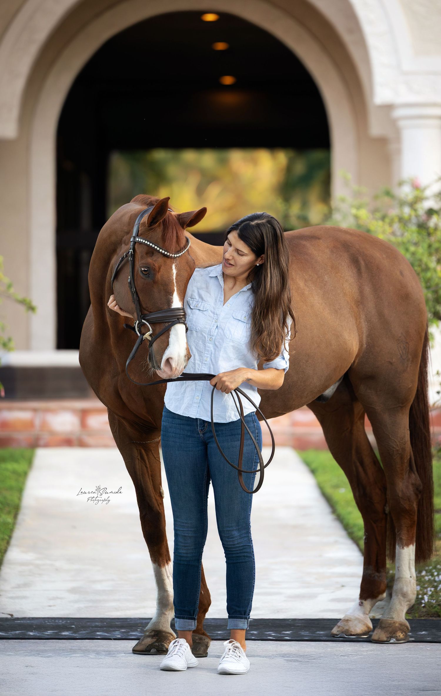 Brown horse stands with handlers by stone archway columns.