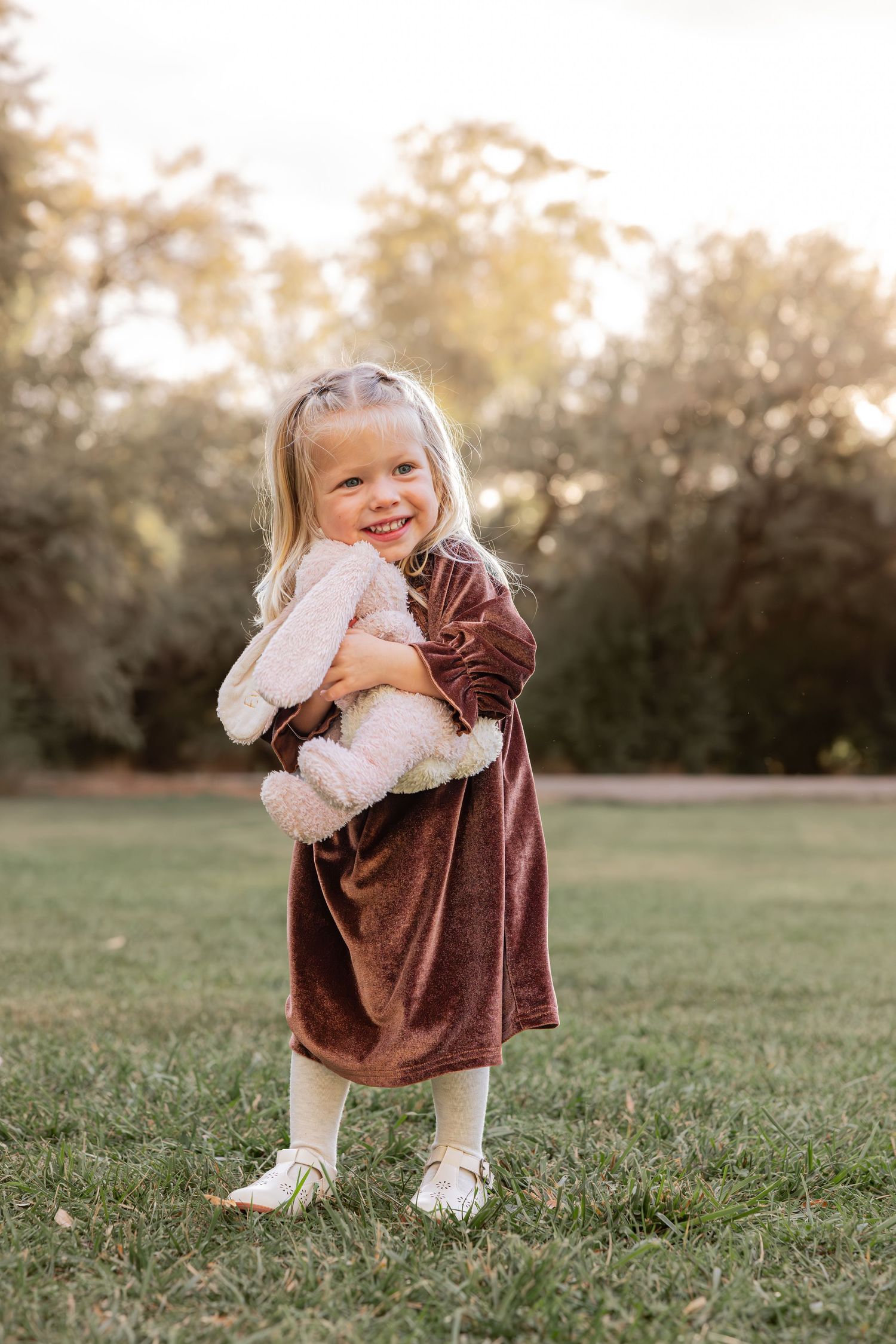 A young child in a velvet dress holds a stuffed toy while smiling outdoors.