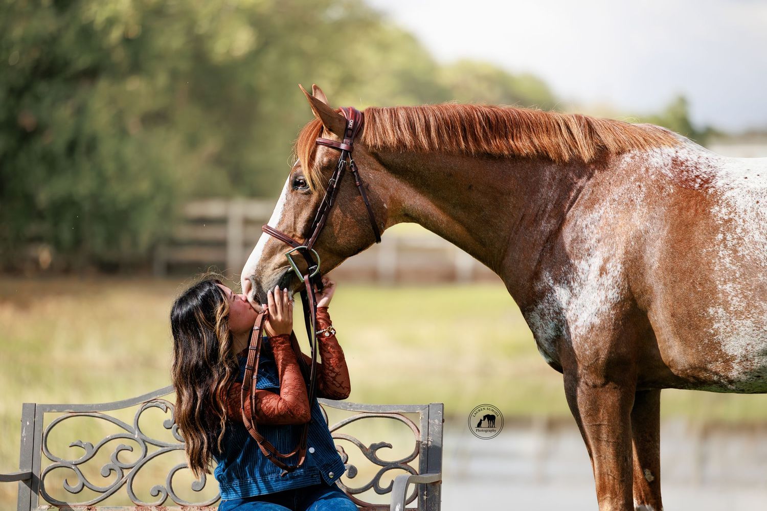 A horse and rider share a tender moment in a rustic outdoor setting with denim and leather attire.