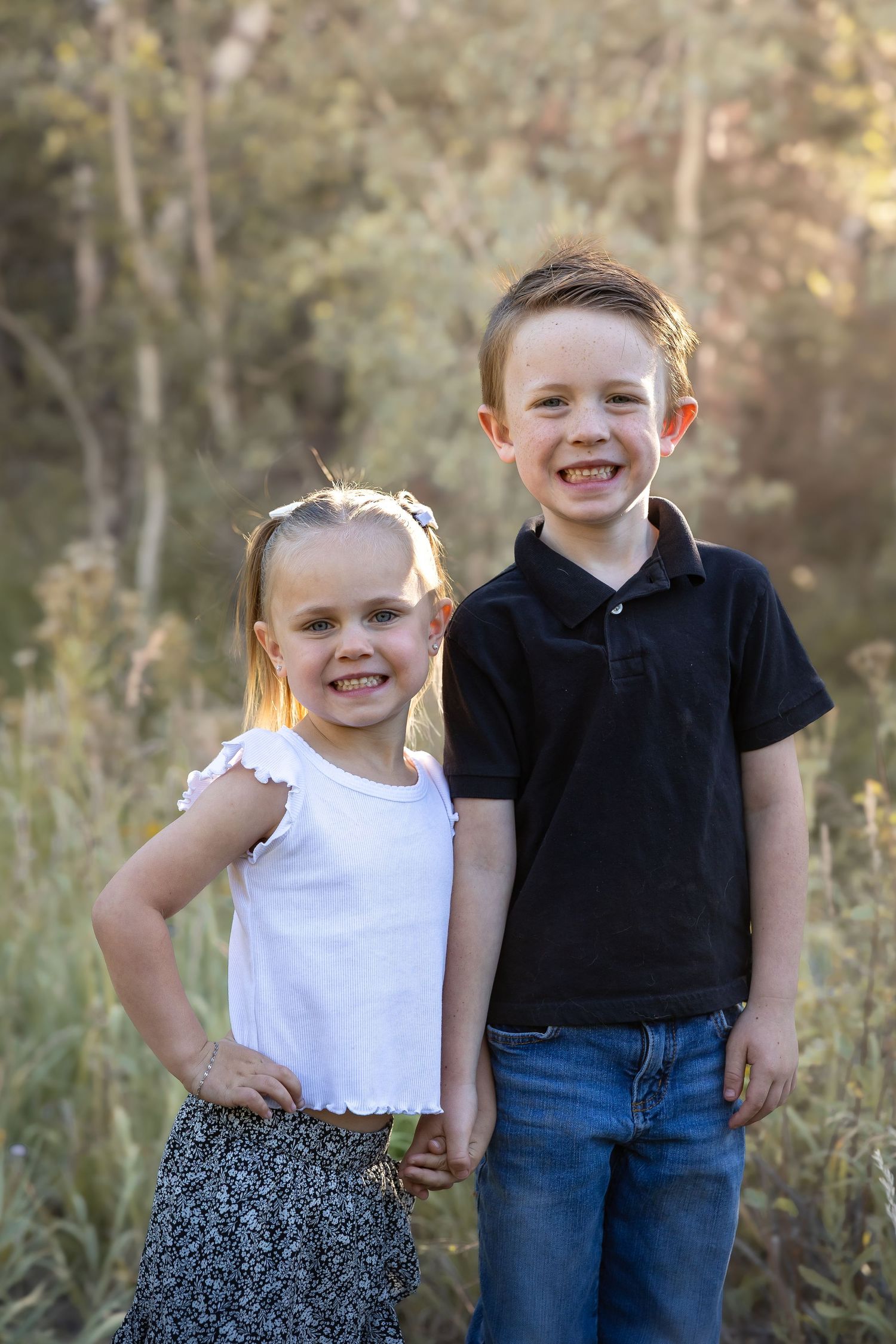 Two children pose together in casual outfits among wild grasses during sunset.