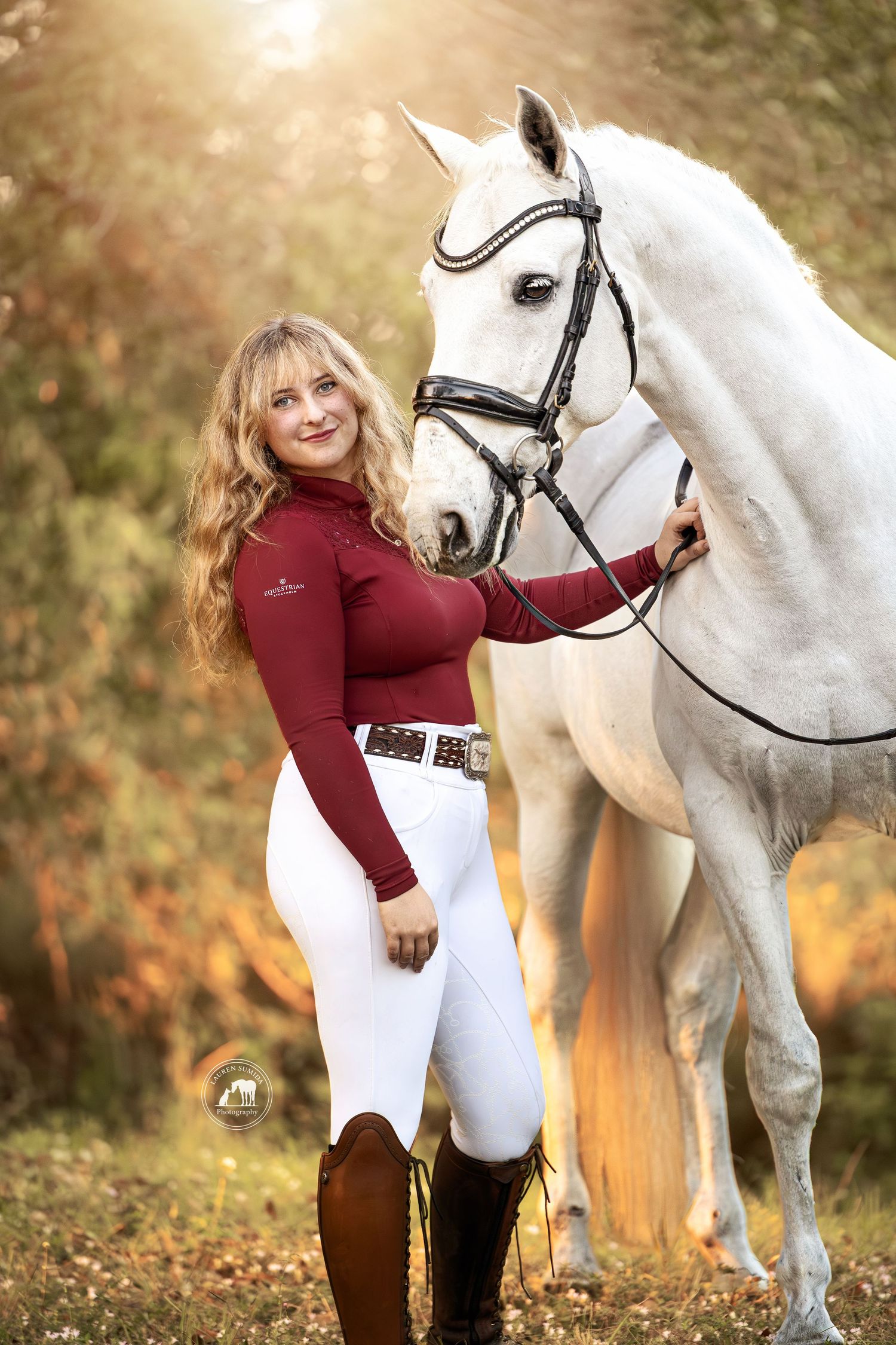 White horse and rider in burgundy top pose in fall setting with golden light.