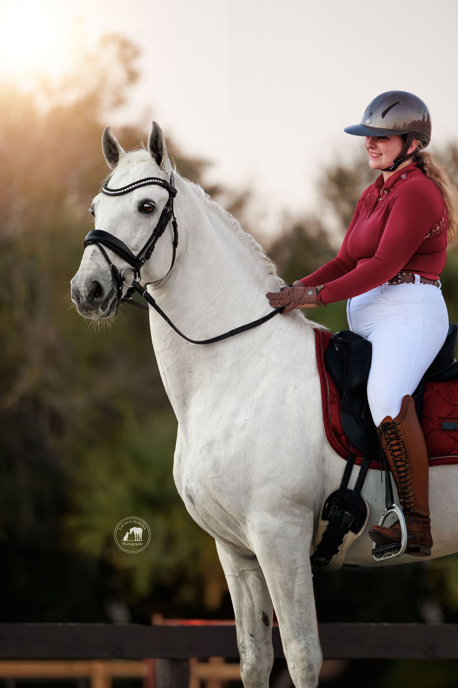 Smiling woman in a burgundy jacket, white breeches and riding helmet sits on a gray horse with a black bridle at sunset.