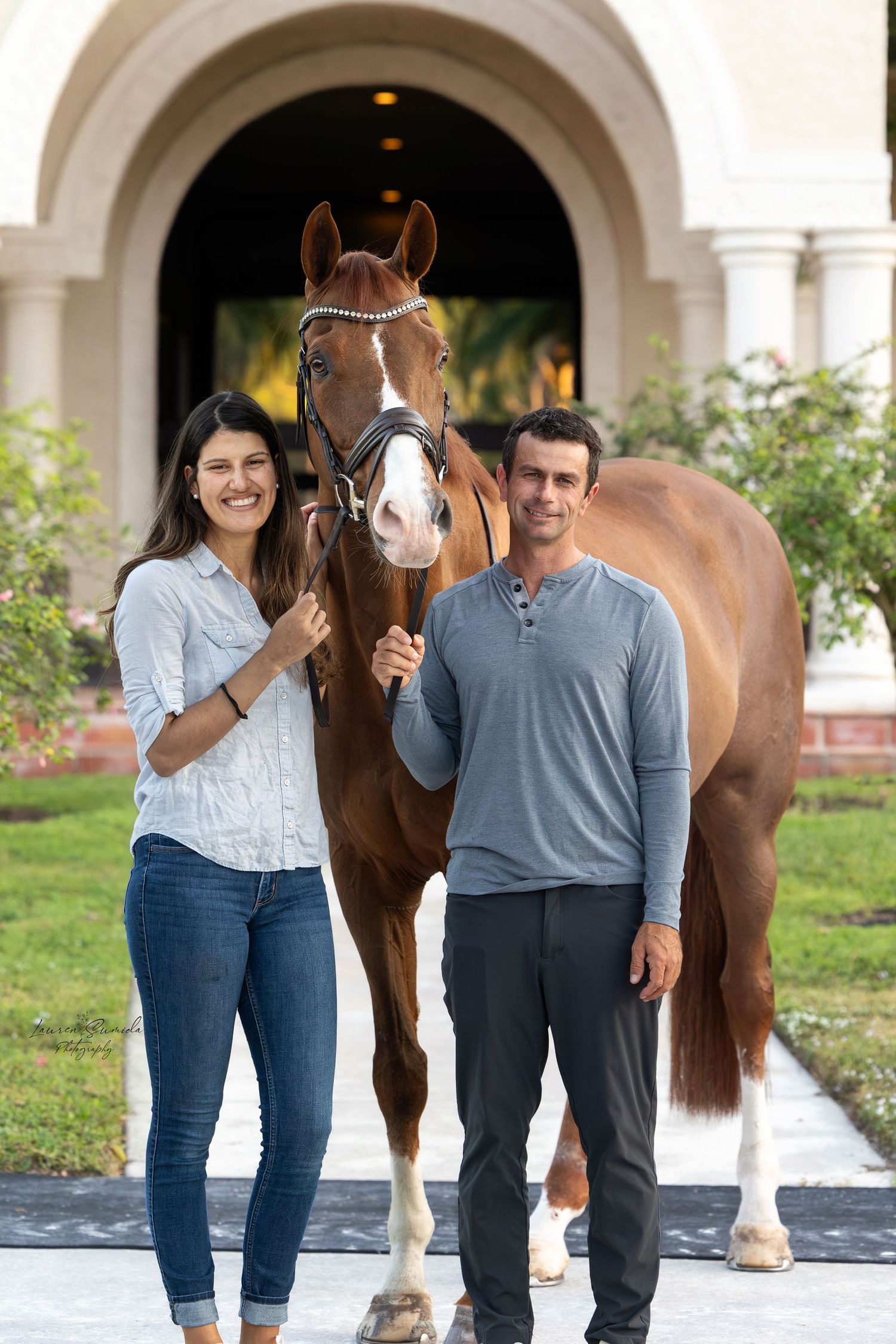 Horse stands between two people in casual attire by an arched stone walkway.
